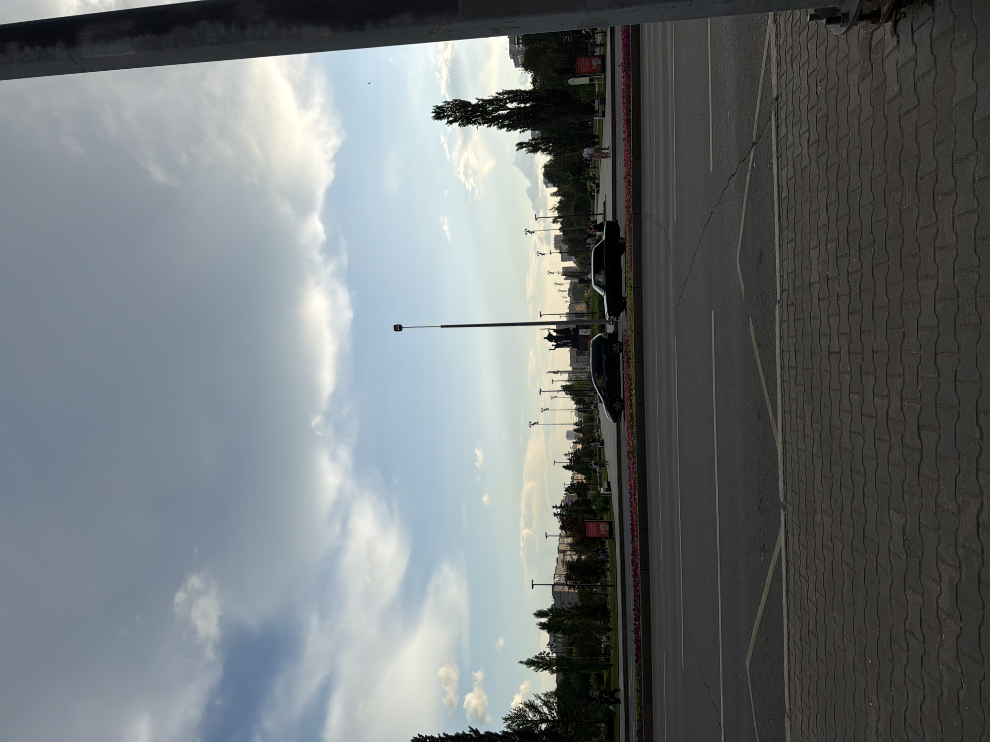 The image depicts a street scene in Perm, Russia, likely in an urban park or public square. In the foreground, a paved pedestrian walkway is visible, leading to a multi-lane road with light traffic. Cars are present on the road, and there is a flowerbed with pink flowers separating the road from the pedestrian area. Beyond the road, a park-like setting unfolds with numerous trees, streetlights, and buildings in the background. Several people are scattered throughout the park, some walking and others standing. The sky above is partly cloudy with a mix of blue sky and white clouds, suggesting it might be late afternoon or early evening. There is also a tall, dark structure on the far right of the image, possibly a utility pole or monument.