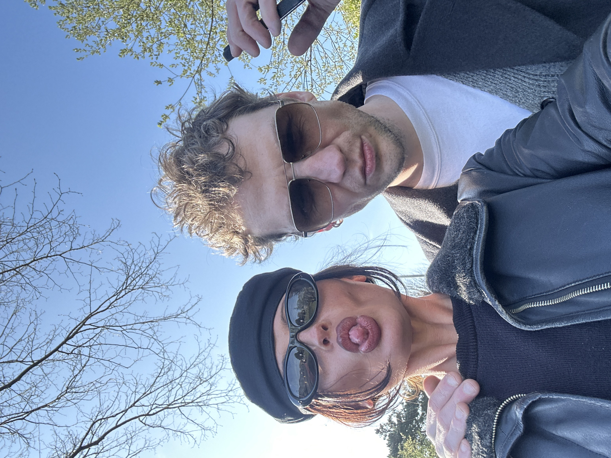 A low-angle photograph depicts two people outdoors. The person on the left wears sunglasses and a black beret, sticking their tongue out. The person on the right wears sunglasses, a white T-shirt, and a jacket. Bare tree branches are visible against a blue sky. Green foliage is visible in the background. The location is the Folkestone and Hythe District, United Kingdom. The picture is taken with a mobile phone.