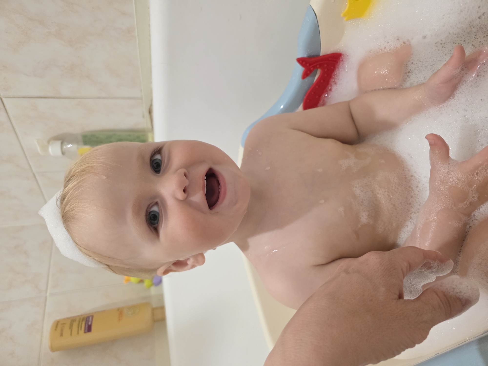 A baby is in a bathtub filled with bubbles, with bubbles also on its head. An adult hand is also visible, interacting with the bubbles. Two bottles sit on a tiled surface next to the tub. A red toy shaped like the number 7 is visible in the tub. No information connects the image to Chaikovsky, Russia.