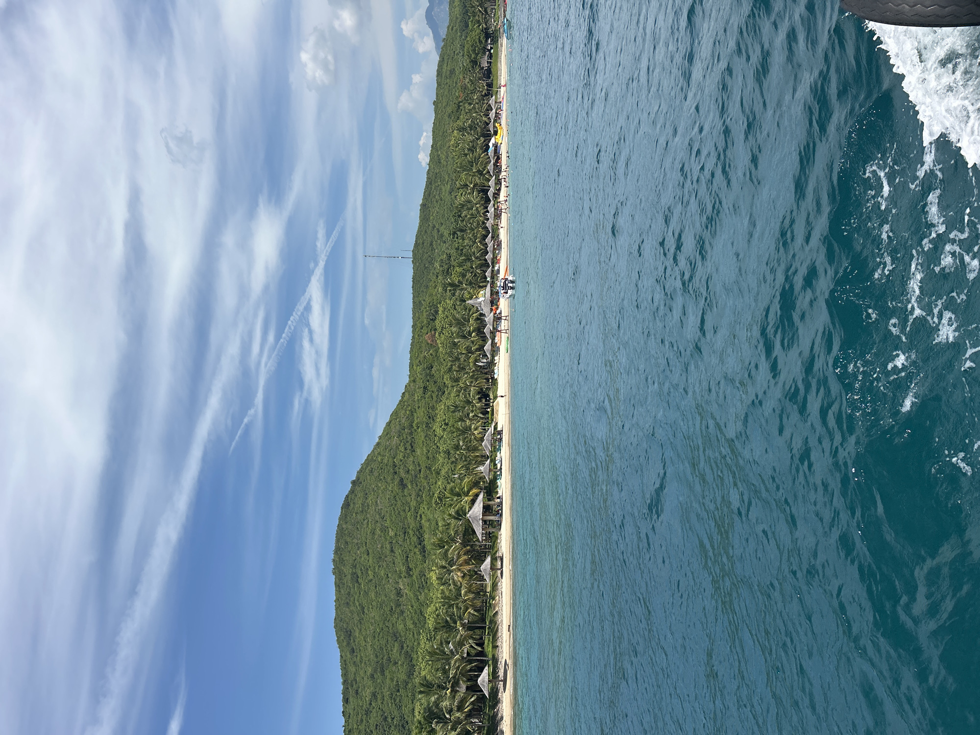 This is a daytime, eye-level, long shot image of a tropical beach resort area in Nha Trang, Vietnam, viewed from the water. The water in the foreground is a deep blue-green, with white foam and spray visible on the right side, indicating the movement of a boat.
The middle ground features a sandy beach lined with palm trees. Several thatched-roof huts and beach umbrellas are scattered along the shoreline, with lounge chairs set up. Small groups of people can be seen relaxing and walking on the beach. A yellow inflatable structure is visible in the water near the shore, and a small white boat is in the water closer to the beach.
The background is dominated by a lush, green, tree-covered mountain that slopes down towards the beach. The sky is a bright blue with scattered white, wispy clouds. A tall antenna or tower is visible on the crest of the mountain in the distance. The overall scene depicts a vibrant and popular tourist destination.