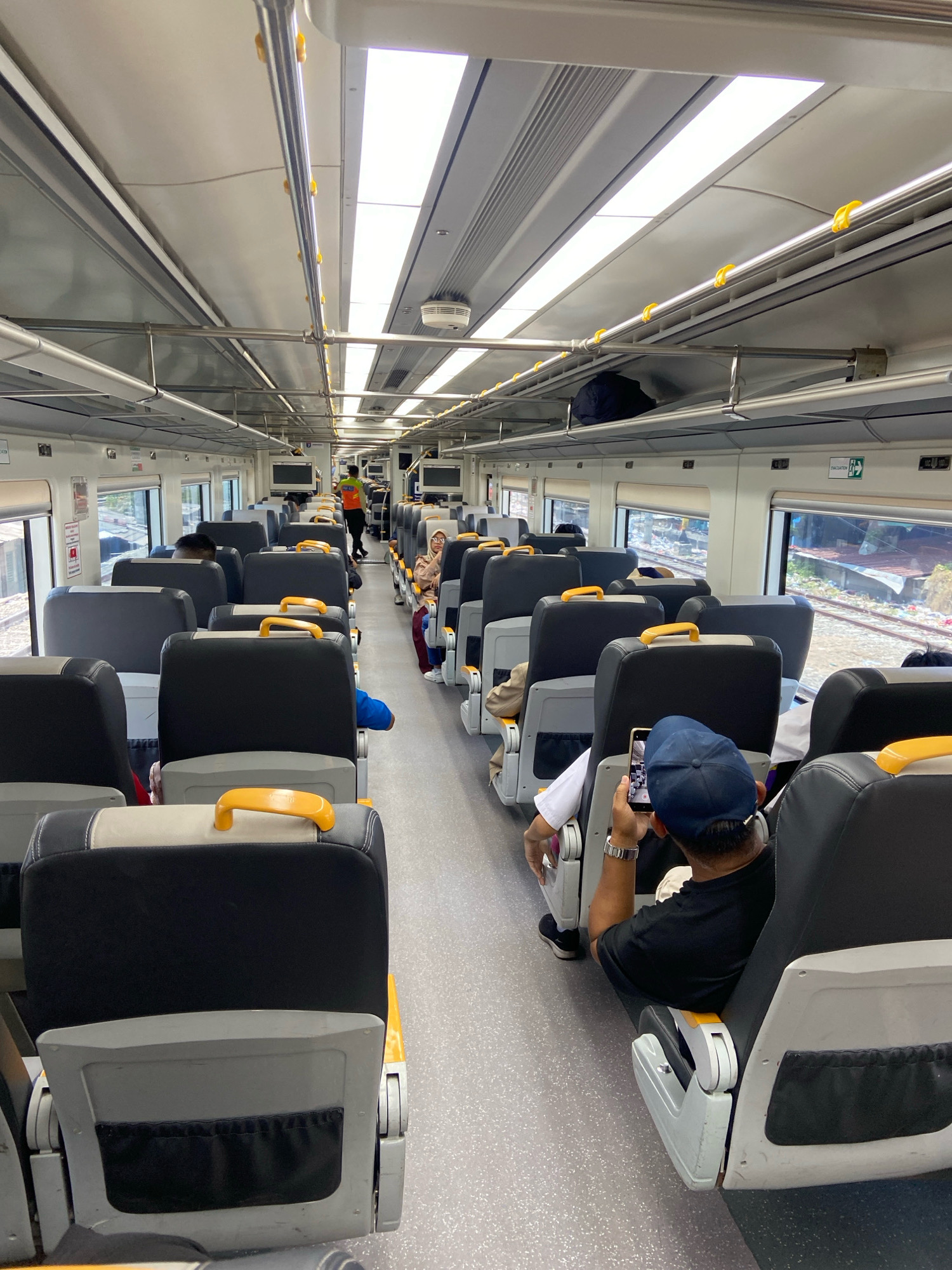 The image shows the interior of a commuter train carriage in North Jakarta, Indonesia. The carriage is modern and well-lit, with rows of grey and black seats on either side of a central aisle. Yellow handles are visible on the backrests of the seats. Passengers are seated, some looking at their phones, while others gaze out the windows. A train attendant in an orange vest is visible walking down the aisle. The train appears to be in motion, as suggested by the blurred scenery outside the windows. The overall impression is one of a typical commute on an Indonesian train.