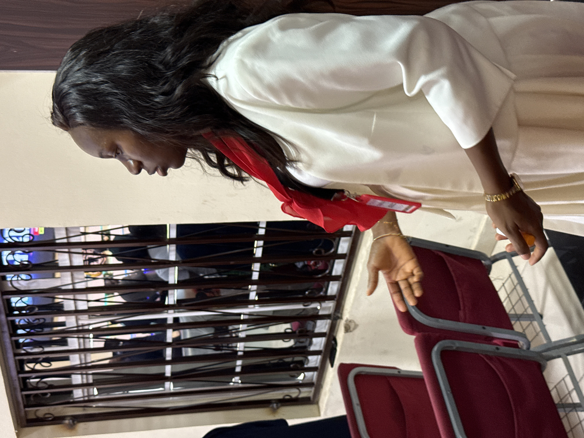 In a brightly lit indoor setting, possibly a church or meeting hall, a woman stands in profile facing left, her dark, wavy hair cascading over her shoulders. She wears a white blazer over a white top and skirt, accented by a vibrant red scarf around her neck and a red lanyard with an identification card. Her right hand is extended, palm open, while her left hand holds a small orange object. In the background, through a barred window, several indistinct figures are visible, and to her left, two rows of maroon chairs with grey metal frames are arranged. The overall environment appears to be clean and well-maintained, suggesting a public or communal space.