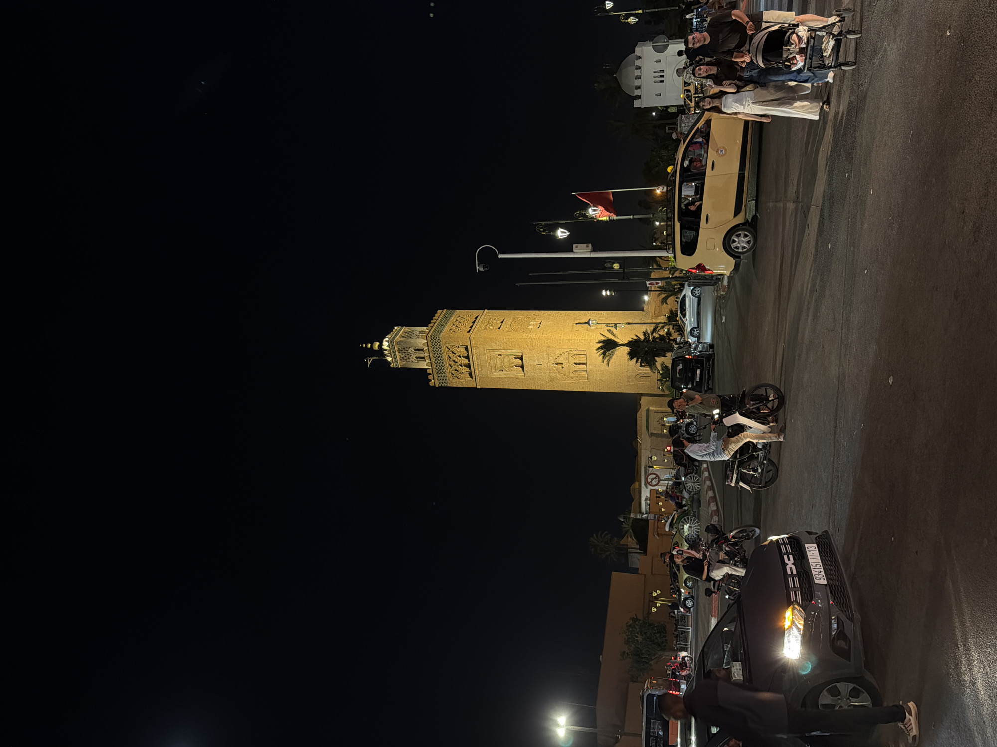 The image depicts a nighttime scene in Marrakech, Morocco, likely in the vicinity of the Koutoubia Mosque. A tall, illuminated minaret dominates the background, casting a warm glow. The foreground features a busy street intersection with several vehicles, including a dark grey car with its headlights on, a yellow taxi, and a bus. People are seen walking and riding motorcycles. A family is crossing the street, with a person pushing a stroller. The atmosphere is lively, with streetlights illuminating the area. The dark night sky above provides a stark contrast to the illuminated structures and vehicles.