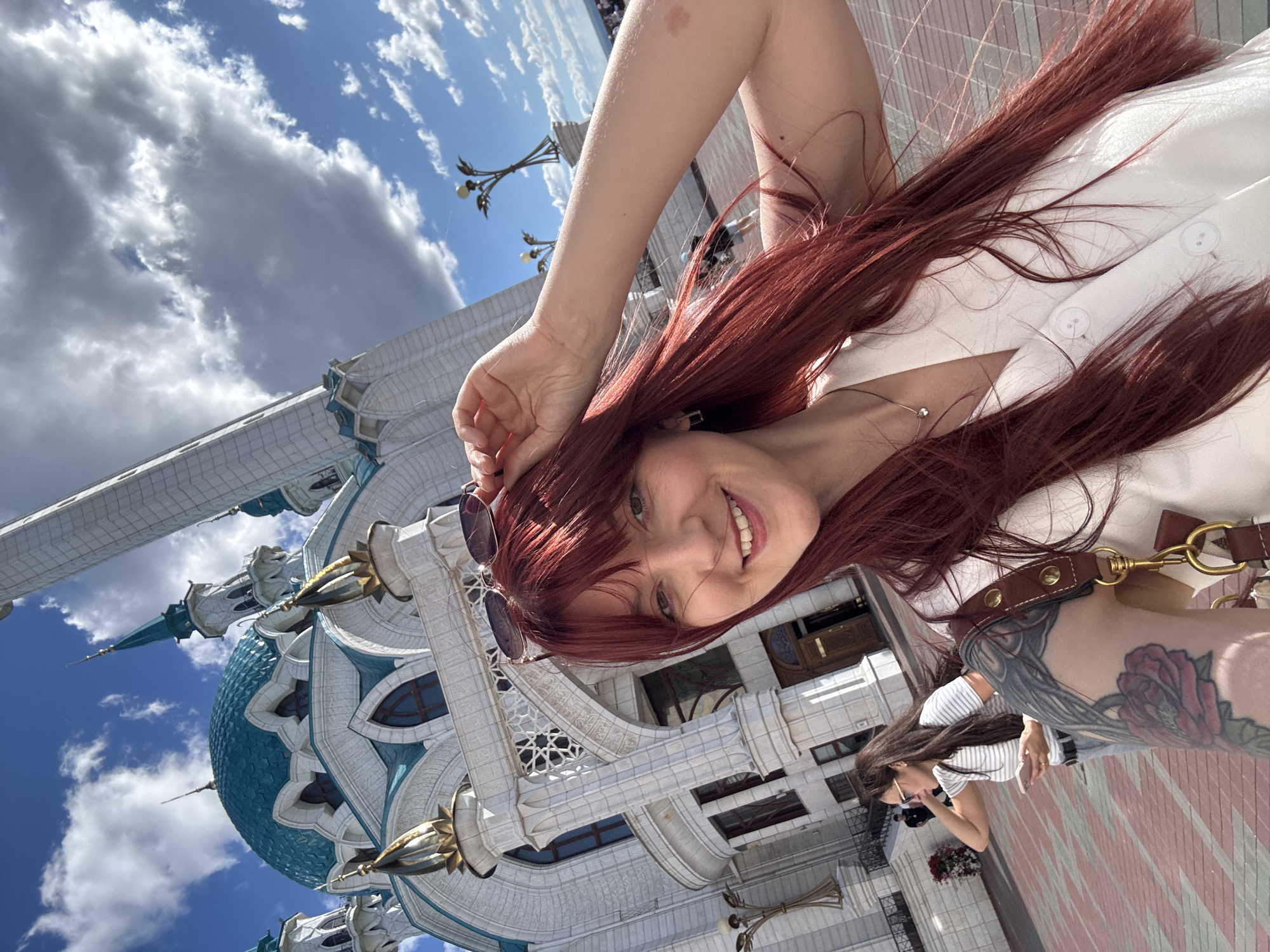 A young woman with long, auburn hair and bangs is taking a selfie outdoors in front of a large, white mosque with a turquoise dome and minarets. She is wearing a white vest with two visible buttons and a silver necklace with a small pendant. Sunglasses are perched on top of her head. Her right arm is raised, and she is touching her sunglasses. A large rose tattoo is visible on her left arm.
In the background, another woman with dark hair, wearing a striped t-shirt and sunglasses, is looking at her phone and walking away from the camera. She is wearing jeans.
The mosque appears to be the Kul Sharif Mosque in Kazan, Russia, characterized by its distinctive architecture, including ornate stonework, gold accents, and vibrant turquoise domes. The sky is blue with scattered white clouds. The ground in front of the mosque is paved with red and grey tiles. Several decorative lampposts are visible in the background, as are other people walking around the grounds. The overall impression is a sunny day at a prominent landmark.