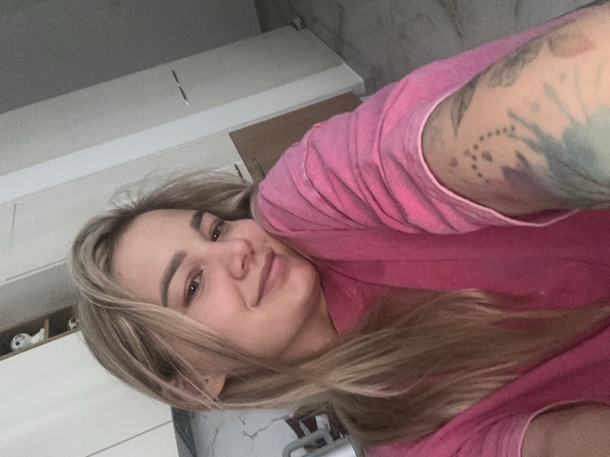 A woman with long, blonde hair and a pink shirt is looking at the camera with a slight smile. Her left arm, adorned with a tattoo, is visible in the upper right corner. The background shows a room with white cabinets and a light-colored wall. There is no indication of Budva, Montenegro.