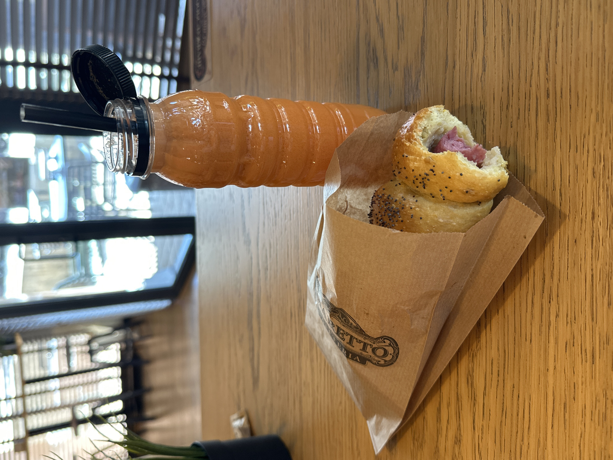 The image shows a close-up view of a wooden table with a bottle of orange juice and a pastry in a paper bag. The bottle of juice is on the right side of the image, with a black lid and a black straw. The pastry, which appears to be a pretzel-like bread topped with poppy seeds and filled with pink meat, is in a brown paper bag on the left side of the image. The paper bag has a circular logo with text on it, which is partially obscured. The background is blurred and shows a restaurant interior with blinds on a window, suggesting a commercial setting. The lighting suggests it is daytime. The location is not specified in the image itself, but it is stated to be in the city of Veliko Tarnovo, Bulgaria. There are no people visible in the image.
