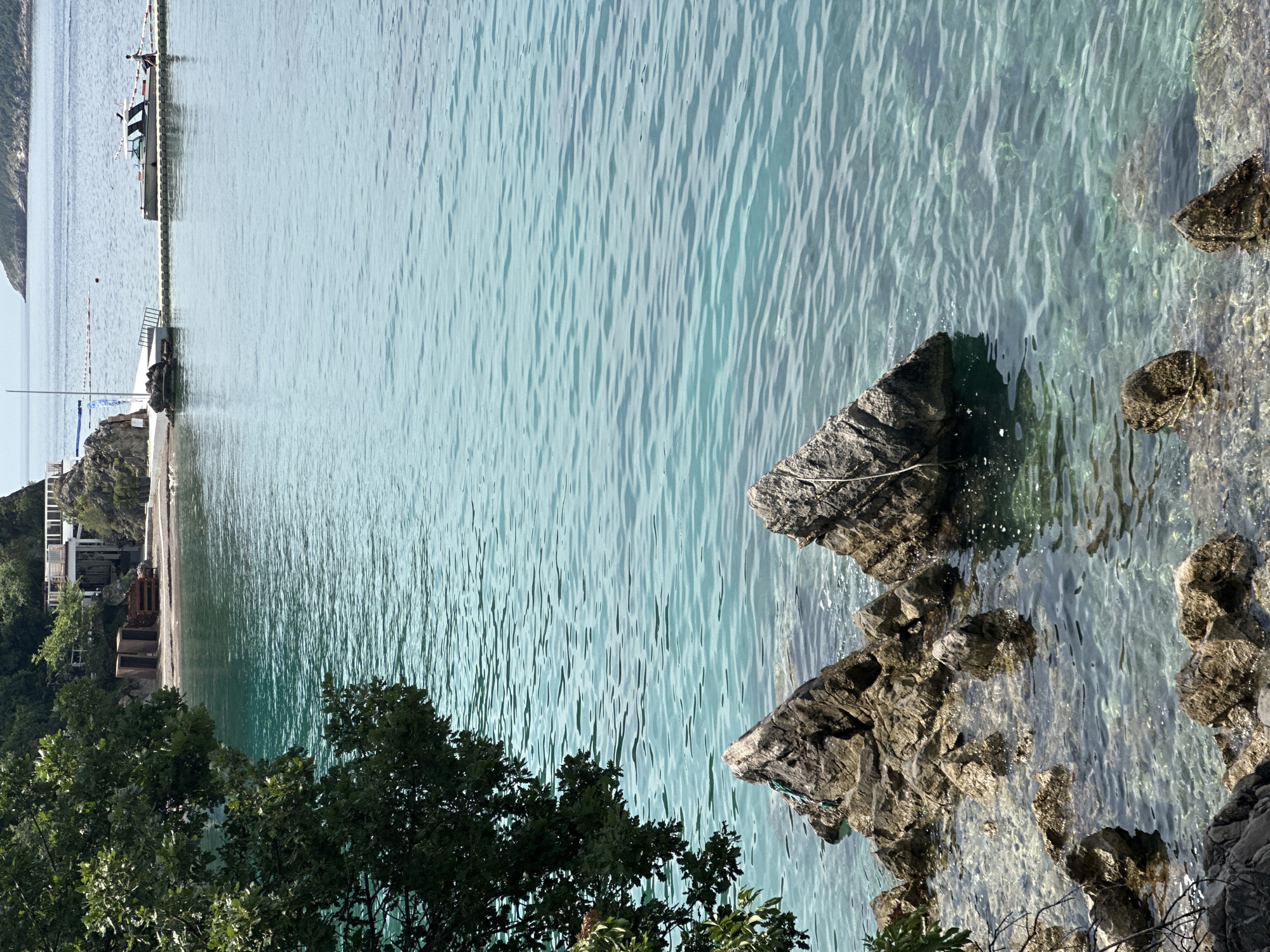 A vertical, eye-level shot captures a picturesque scene of the Adriatic Sea off the coast of Budva, Montenegro. In the foreground, rough, grey rocks jut out of the crystal-clear, turquoise water, some partially submerged, revealing the rocky seabed beneath. A dark green tree branch, laden with leaves, extends into the frame from the left, partially obscuring the view of the shoreline.
In the mid-ground, a sandy beach is visible, backed by a rocky outcrop. On the beach, there are several weathered wooden structures that appear to be changing rooms or storage units, and a pile of wooden planks. A white building with balconies and railings is perched on the rocky hillside above the beach.
Further out, a long, modular pontoon pier extends into the sea, with a white boat docked at its end. A person in dark clothing is walking along the pier towards the boat. Red and white buoys mark a swimming area further out in the bay. In the distance, the hazy outline of a mountainous island completes the serene coastal panorama. The water is calm, with gentle ripples reflecting the bright, overcast sky.