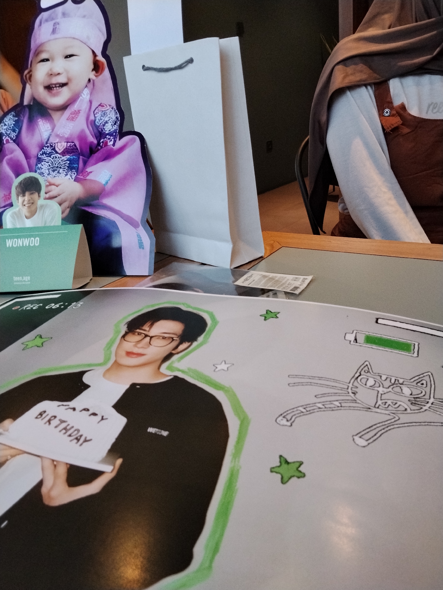 In a cafe in Serang, Indonesia, a cardboard cutout of a smiling baby in a traditional Korean hanbok outfit stands to the left of a white paper shopping bag with a gray drawstring. To the right, a person wearing a brown vest over a light gray sweatshirt and a brown headscarf is partially visible. In the foreground, a large, printed poster displays a photo of a young man with glasses, wearing a black jacket over a white shirt and holding a small white cake with "HAPPY BIRTHDAY" written on it. A stylized drawing of a cat and green stars are also present on the poster. A smaller cutout of the young man in a white shirt is attached to a green cardboard stand that reads "WONWOO" and "teen,age". The scene appears to be set on a table.