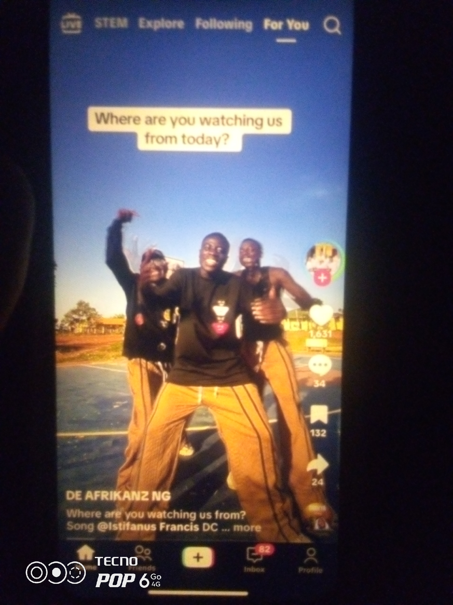 This is a photograph of a smartphone screen displaying a social media video. The video features three young Black men, two of whom are prominently in the foreground, smiling and gesturing. They are wearing black t-shirts and orange pants. The background appears to be an outdoor setting with a basketball court and some distant buildings and trees. The screen also shows text overlays, including "Where are you watching us from today?", "DE AFRIKANZ NG", and "Song @Istifanus Francis DC ... more". There are also icons for likes, comments, shares, and other social media features visible on the right side of the screen. The bottom of the screen displays the TECNO POP 6 4G logo. The photograph is taken in low light conditions, likely indoors at night, with the smartphone screen being the primary light source. The specific location of Howayi, Nigeria, is not discernible from the image itself, as it is a photograph of a screen, not the actual environment.