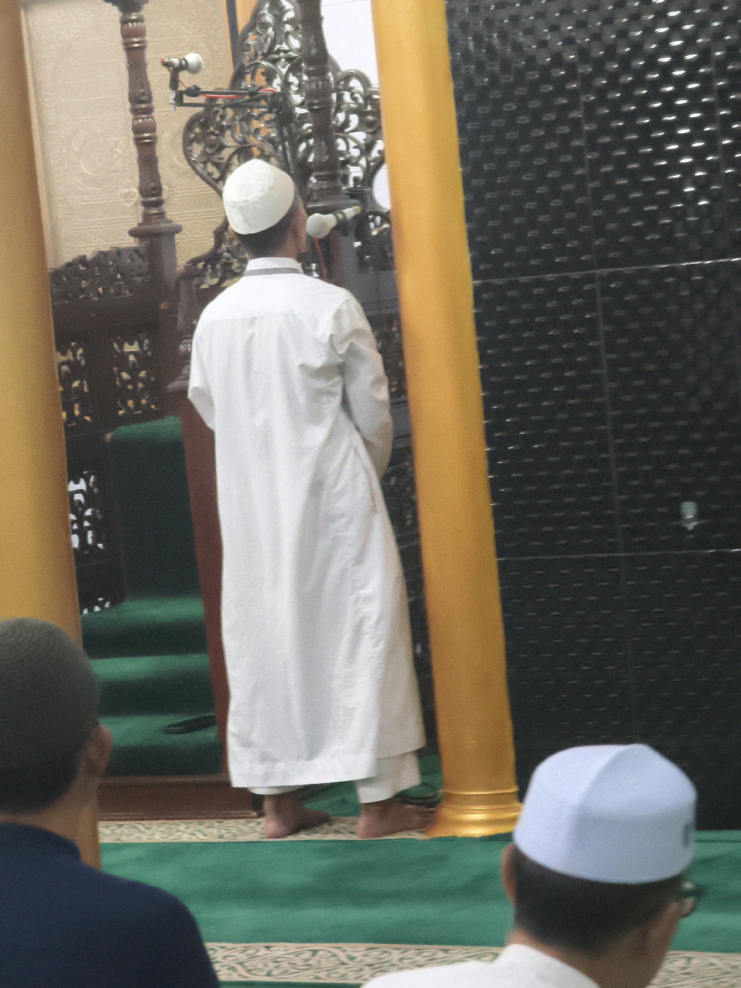 In Samarinda, Indonesia, inside a mosque, a person wearing a white peci (cap) and a white robe stands facing away from the camera, with their back to a decorative wooden structure that appears to be a pulpit or minbar. Another person, also wearing a white peci, is visible in the foreground, facing towards the back of the mosque. The environment features green carpeting with a patterned border, and a large, dark, textured wall is visible in the background. A yellow pillar is positioned to the right of the person in the white robe.
