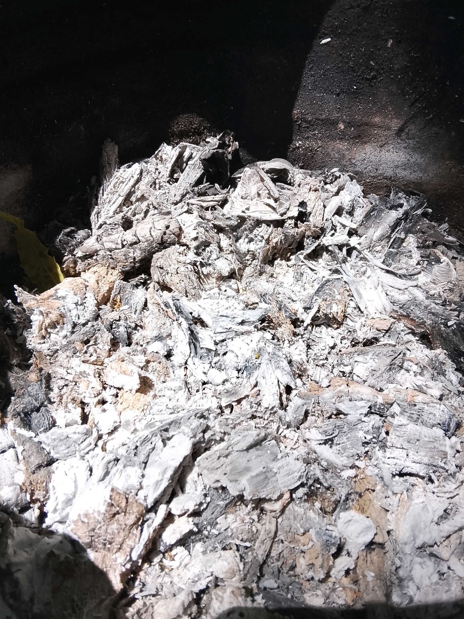 The image shows a pile of ash and burnt material in what appears to be the bottom of a fire pit or grill. The ash is predominantly white and gray, with some darker, charred pieces mixed in. The texture is rough and uneven, with many fragmented pieces. The surrounding area is dark, suggesting it is a shaded or enclosed space. There is a hint of a yellow object on the left edge of the frame, partially obscured by the ash. The image is a close-up view, focusing on the texture and composition of the ash. There are no people or animals visible in the image, and no specific objects other than the burnt debris. The environment is difficult to determine precisely, but the context suggests an outdoor setting like a garden or patio, potentially in Colombo, Sri Lanka, given the provided location hint, though this cannot be confirmed solely from the visual information.