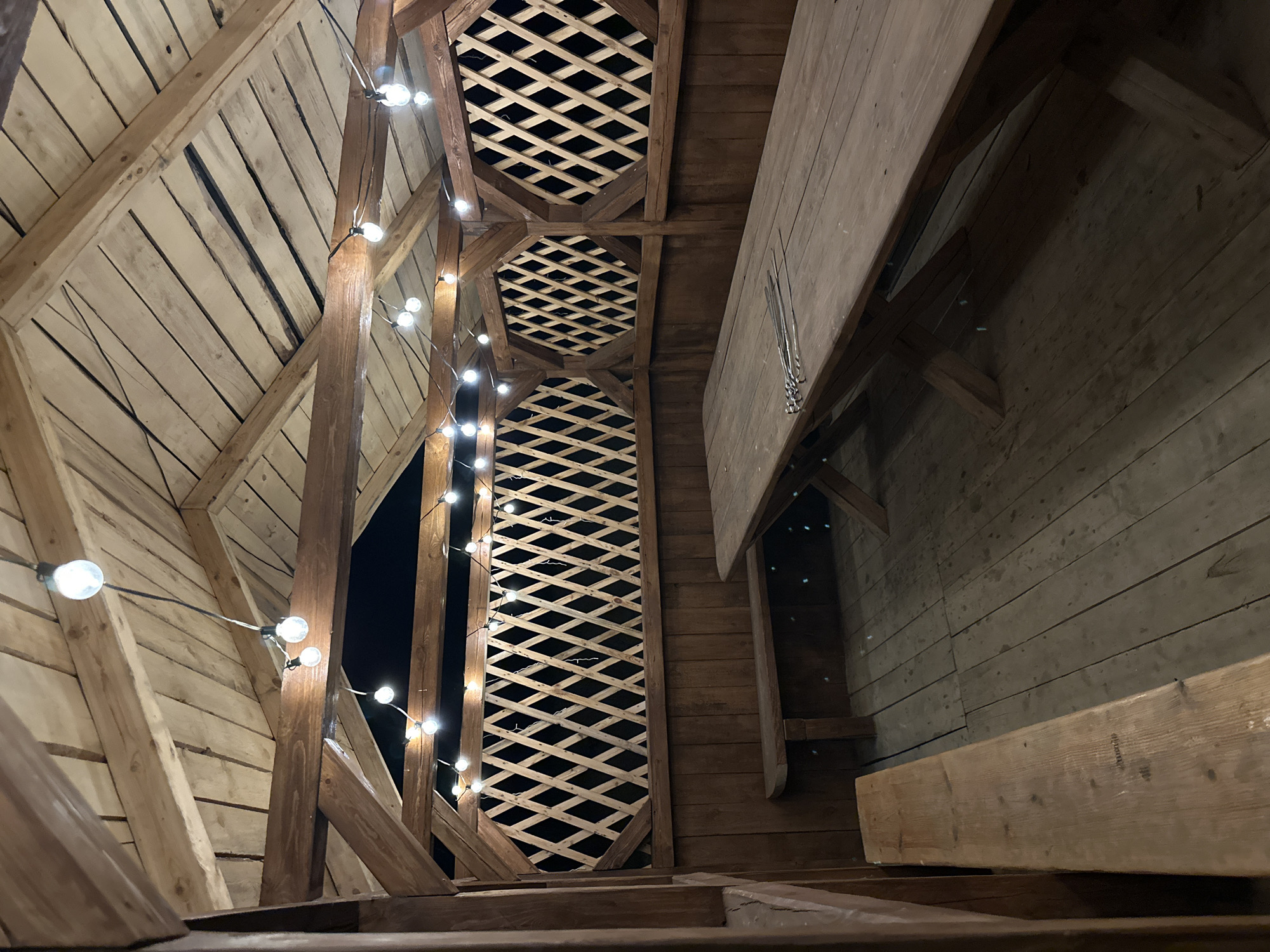 The photograph depicts an interior space with a wooden architectural structure, illuminated by string lights. The structure appears to be an outdoor or semi-outdoor pavilion or gazebo. The walls and ceiling are constructed from wooden planks, with some sections featuring a lattice design. A wooden table is visible on the right side of the frame, and a bench or ledge is situated below. The setting is dark, with the background showing glimpses of the night sky through the lattice. The location is unspecified.