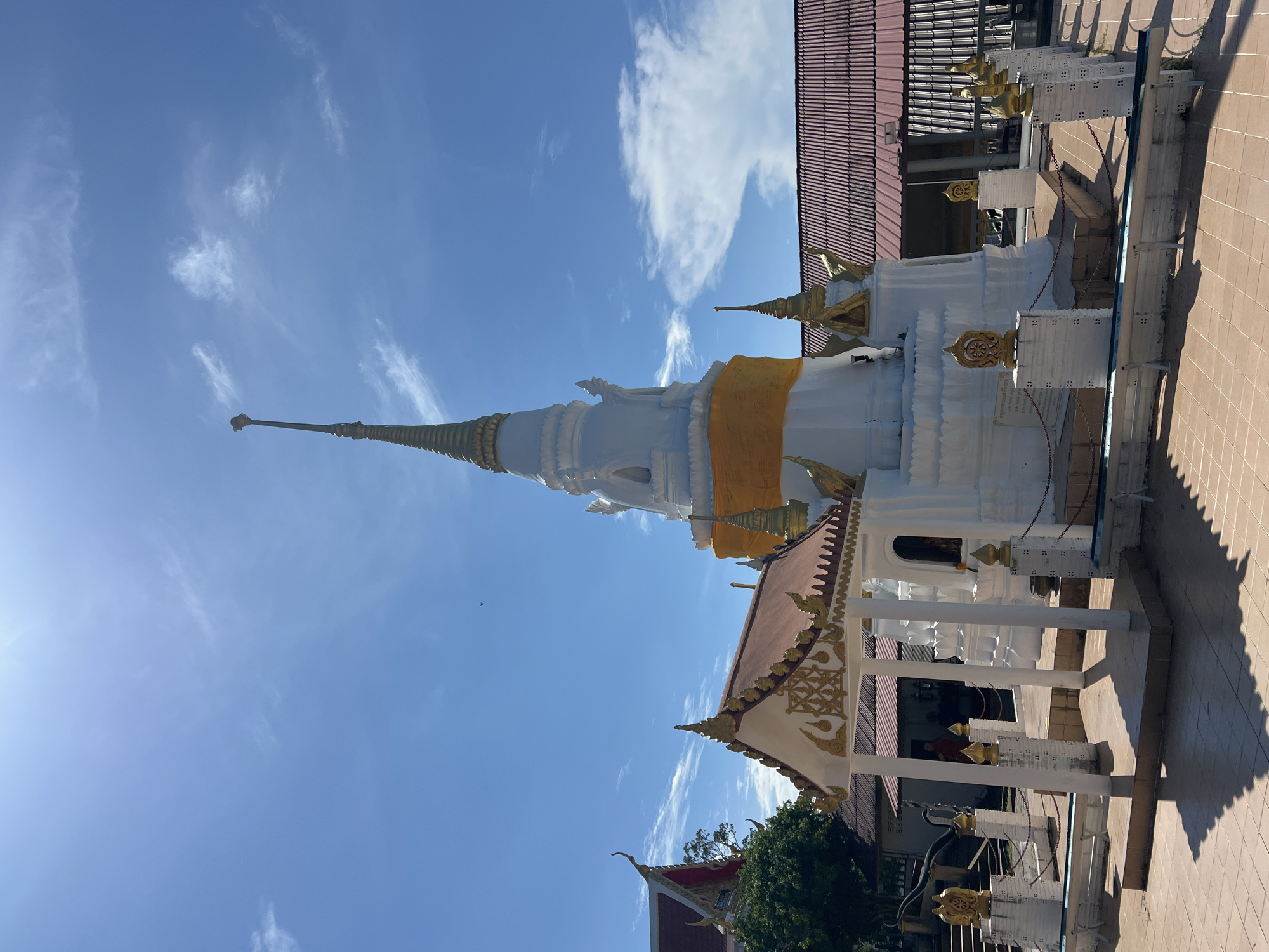 This image depicts a white stupa in the city of Baan Hua Thanon, Thailand, under a bright blue sky with scattered white clouds. The stupa, adorned with a vibrant orange sash, stands tall and conical, topped with a slender spire. In front of the stupa is a covered structure with a red roof and ornate golden trim, supported by white pillars. The area surrounding the stupa is paved with light brown tiles, and a low white wall with golden finials and decorative elements encloses the sacred site. Behind the stupa, a building with a red tiled roof is visible. The scene appears to be a religious or cultural site, possibly a temple, given the architectural style. The lighting suggests it is daytime, with shadows cast by the structures. No people are clearly visible, though a figure in orange, potentially a monk, can be seen in the background through an opening in a building.