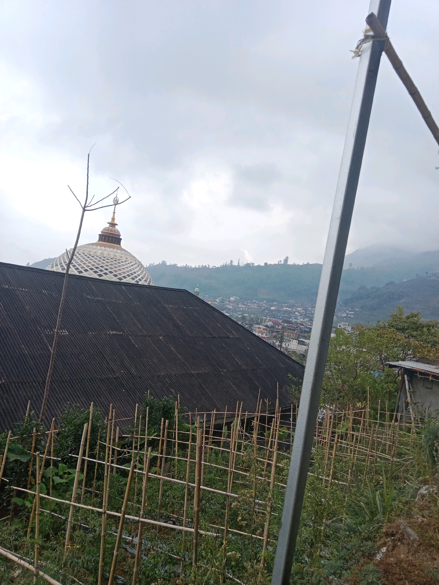 The image captures a view of Surengede, Indonesia, on an overcast day. In the foreground, a row of bamboo supports for a garden is visible, with lush green plants growing behind it. To the left, the dark, corrugated roof of a building dominates the mid-ground. Above this roof, a distinctive golden dome with an intricate patterned surface rises, topped with a spire. A thin, leafless tree branch extends upwards in front of the dome. To the right of the main building's roof, a metal pole stands diagonally, partially obscuring the view. In the background, a sprawling town nestles on a hillside covered in trees and vegetation. The distant mountains are shrouded in mist. A small, light-colored building with a sloping roof and a ladder leaning against its side is situated in the lower right portion of the frame. A very small mosque minaret is visible in the far background on the right.