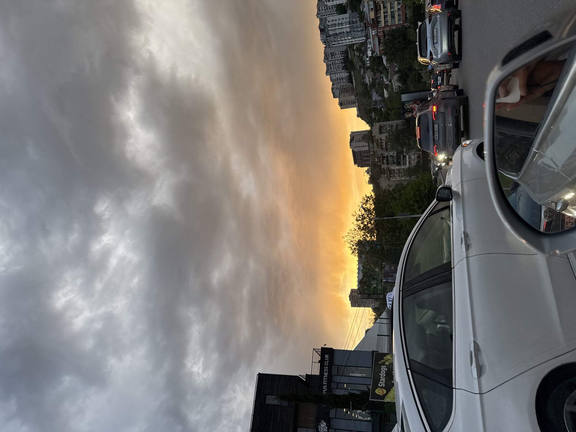 The image shows a street in Tbilisi, Georgia, at dusk or dawn. The sky is divided, with dark, overcast clouds on the left and a vibrant orange-yellow glow on the right, potentially indicating a sunrise or sunset. In the foreground, a white car is visible, partially obstructing the view. A side mirror is also prominently featured, reflecting part of the street scene behind the vehicle. A line of cars is present, suggesting a traffic jam or slow-moving traffic. In the midground, buildings, likely apartments or commercial structures, rise along the hillside. On the bottom left, a building with the words "PUS FITNESS CLUB" is visible. Additionally, a sign indicating "Stardogs" is present. The presence of taxis suggests that the location is an urban area.