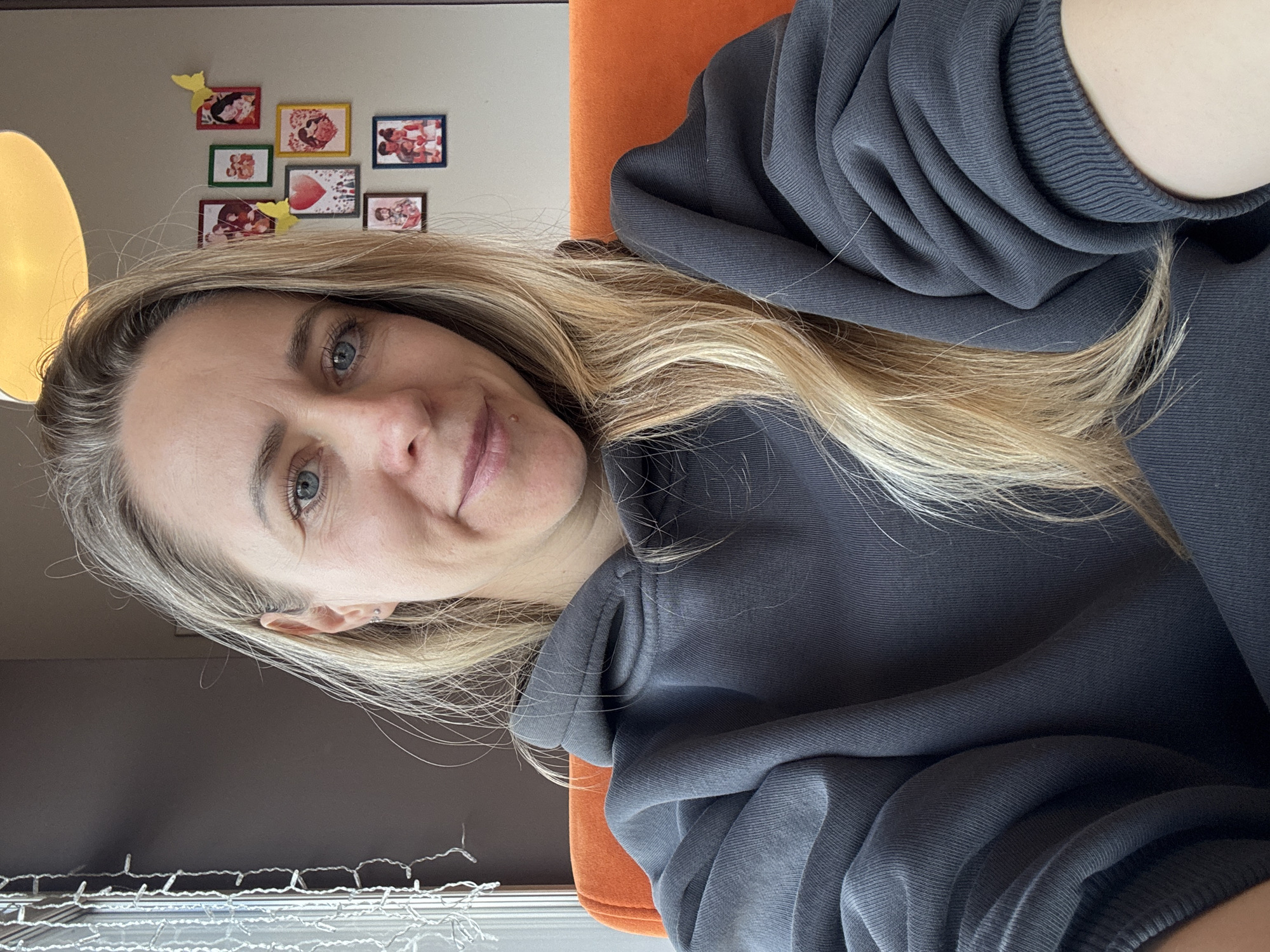 A woman with blonde hair and blue eyes is wearing a dark gray hoodie and sitting in an orange chair. She is indoors, and in the background, there is a wall with several small framed pictures hanging on it. There is also a lamp partially visible in the upper left corner of the image and a string of lights near a window in the bottom left corner. The location of the image is not specified, and there is nothing in the picture to indicate that it was taken in Chaikovsky, Russia.