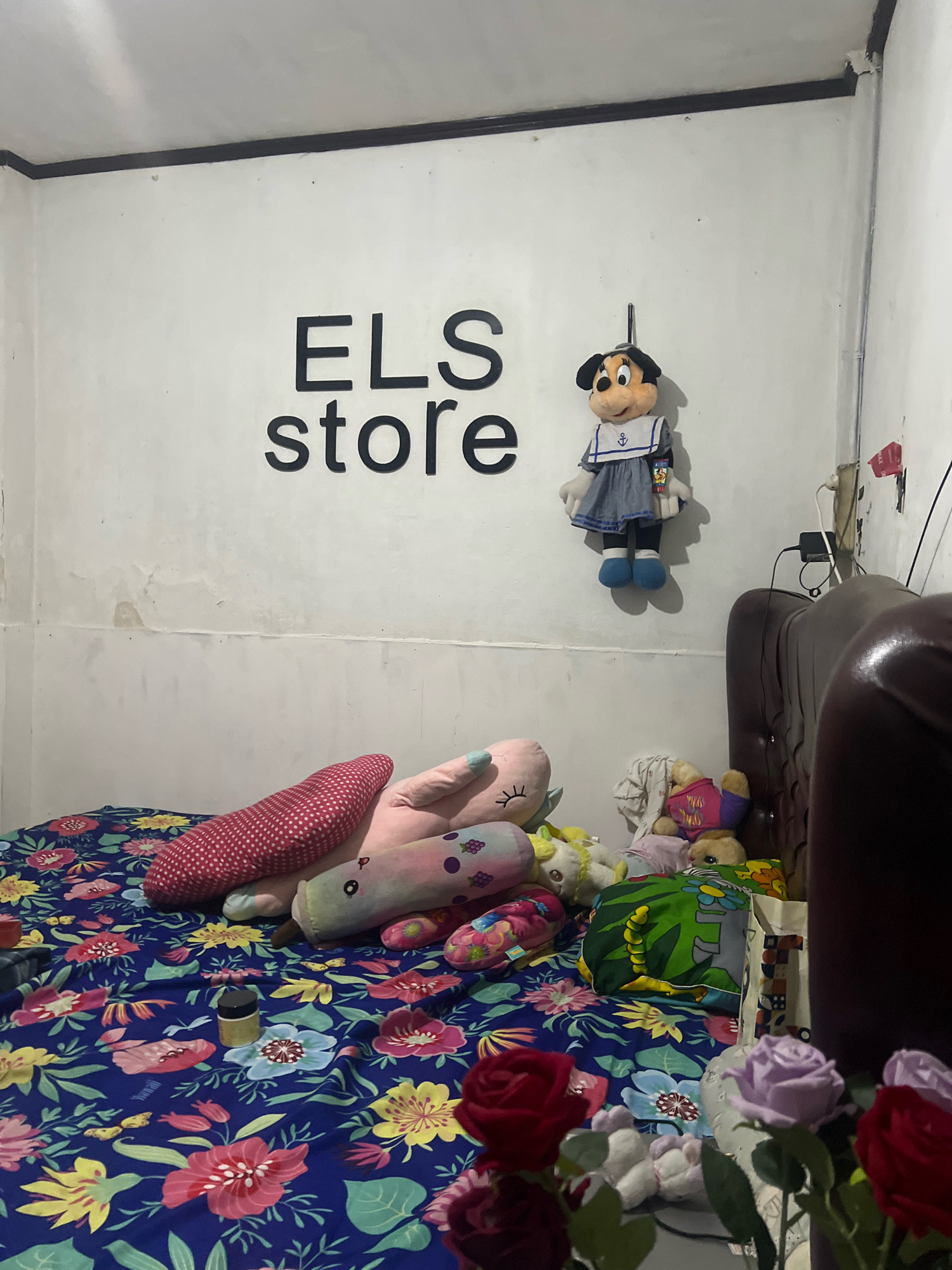 The image depicts an indoor scene, likely a bedroom, filled with stuffed animals and soft furnishings. The room is dimly lit, with a white wall serving as the backdrop. A prominent feature on the wall is the black text "ELS store" written in large, bold letters. To the right of the text, a Minnie Mouse doll dressed in a sailor outfit is hung.
The bed, covered with a vibrant floral blue sheet, is laden with various stuffed animals. These include a pink polka-dotted cylindrical pillow, a long pink stuffed toy with closed eyes, and other smaller plush toys. Some of these are partially obscured by others. A small, dark-capped bottle sits on the bed.
To the right side of the image, a portion of a dark brown, possibly leather, sofa or headboard is visible. Cables and a black adapter are plugged into the wall behind the Minnie Mouse doll, suggesting the presence of electrical outlets and devices. In the foreground, artificial flowers, including red and purple roses, are arranged, adding a touch of color to the scene.
The overall impression is that of a child's room or a space decorated with a playful and cozy aesthetic. The location is specified as Kecamatan Pati, Indonesia, though no specific landmarks or contextual clues within the image directly confirm this geographical placement.