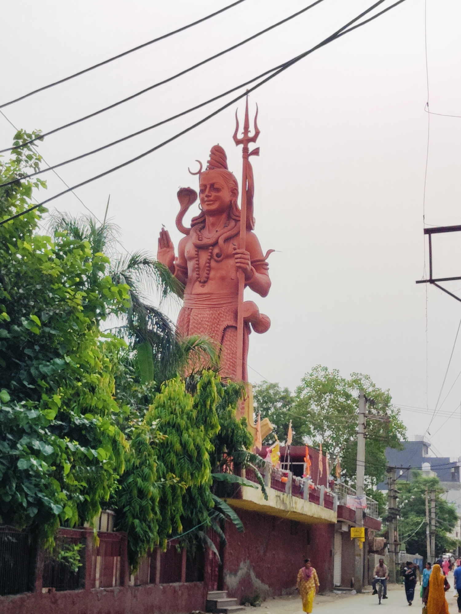 The image shows a tall, reddish-brown statue of the Hindu deity Shiva, holding a trident in his right hand and making a gesture with his left. A cobra is depicted coiled around his neck and arm. The statue is located in Gurgaon, India, and is situated on a raised platform with a building behind it, adorned with prayer flags. In the foreground and to the left of the statue, lush green trees and foliage are visible. The scene is set along a street where several people are walking and a person is riding a bicycle. The sky is overcast. The statue is a prominent landmark in the area.