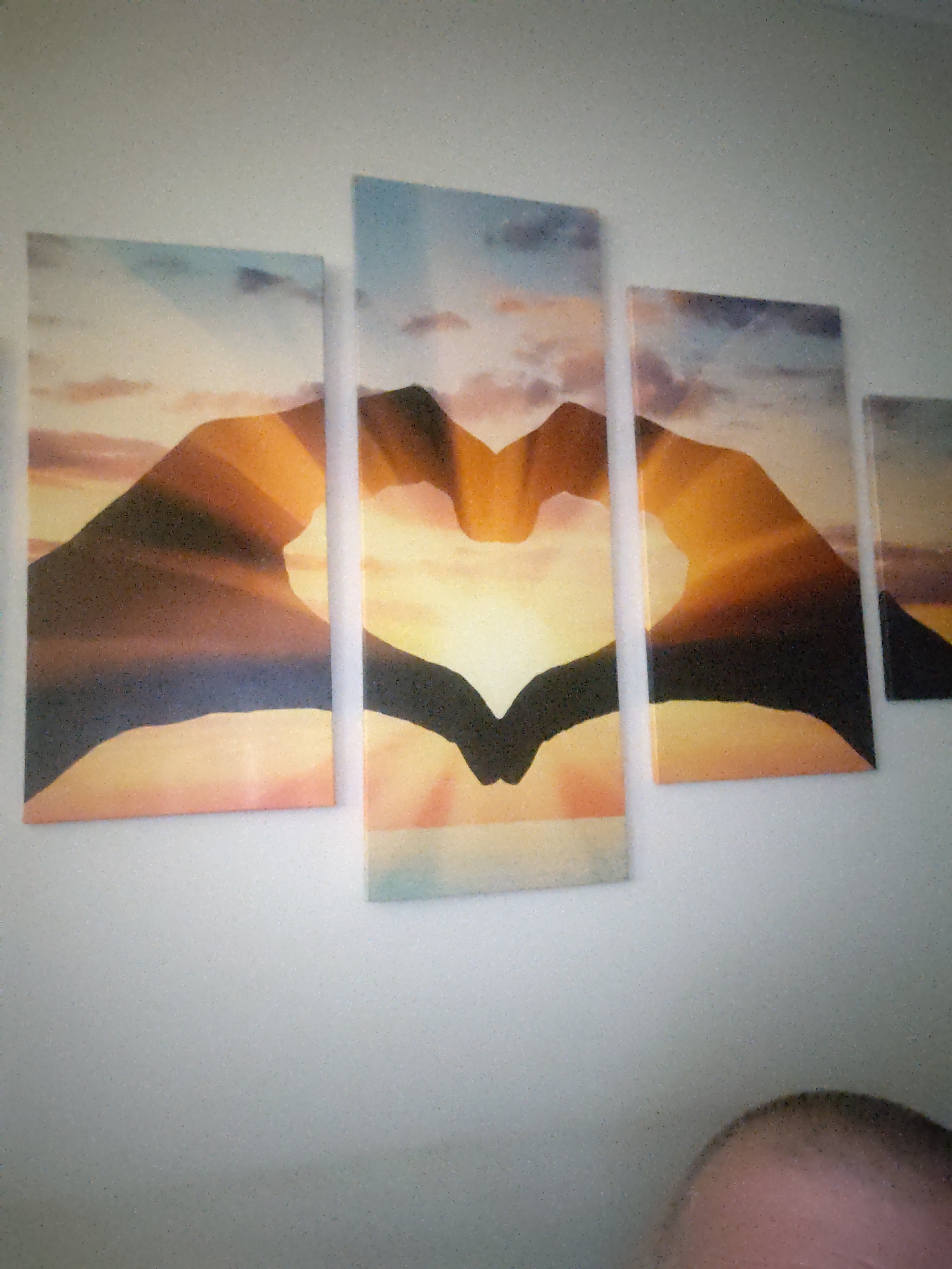 The image displays a five-panel wall art installation horizontally mounted on a light-colored, plain wall in an indoor setting in Huntingdonshire, United Kingdom. The artwork depicts a continuous scene featuring the silhouette of two human hands forming a heart shape against a vibrant sunset or sunrise background. A bright, glowing light emanates from the center of the heart, radiating into warm hues of orange, yellow, and reddish-pink, which transition to cooler blues and grays at the top, indicative of a sky with scattered clouds. The hands are rendered in dark silhouette, with fingers meeting to form the top curve of the heart and thumbs connecting at its lower point. In the lower right corner of the photograph, the top of a human head, including the right ear and a portion of the right shoulder, is partially visible; the hair appears short and dark. The entire photographic image exhibits a fine-grained noise or texture.