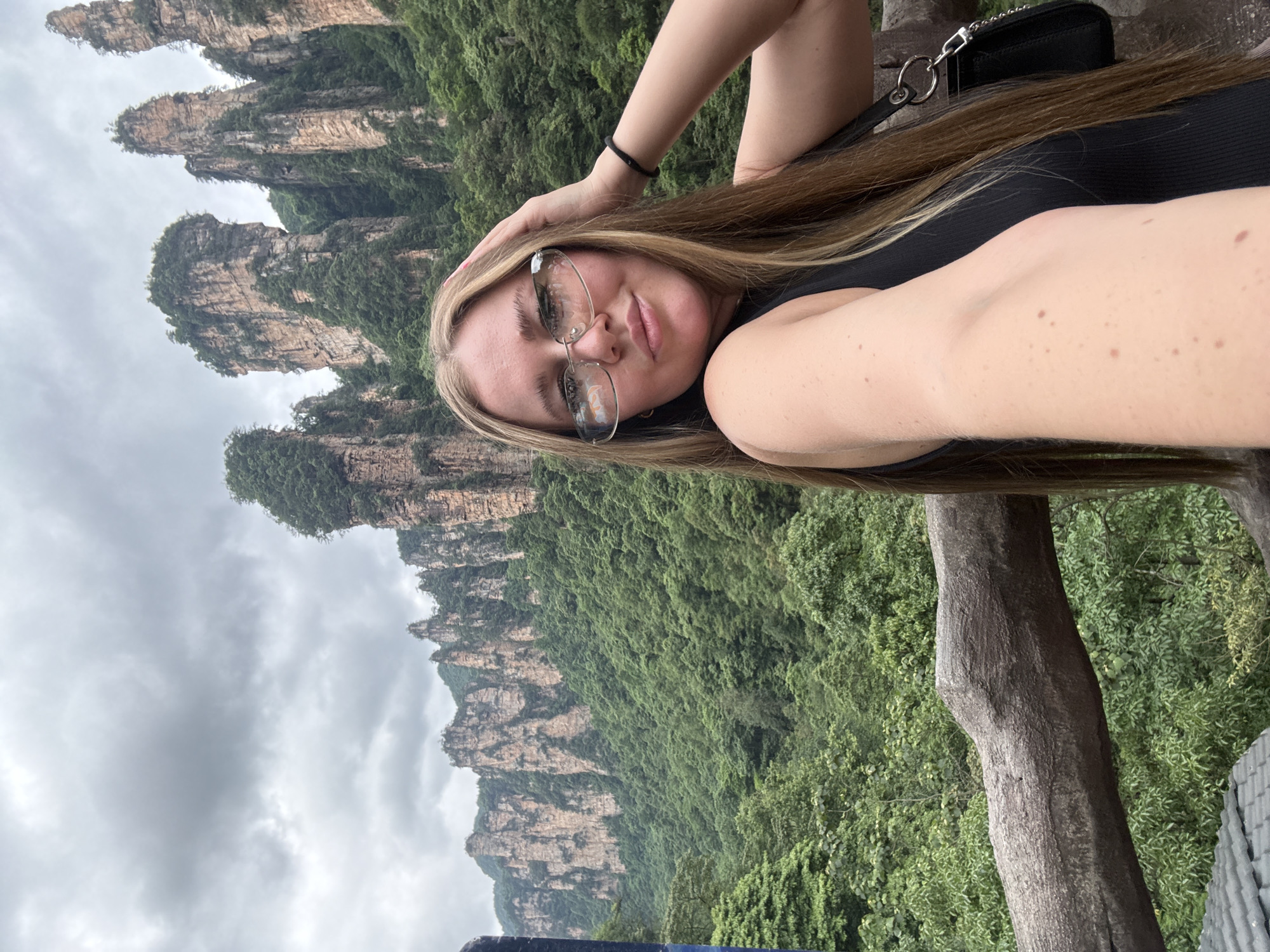 A young woman is photographed in the foreground, with the scenic mountains of Luoguta, China, serving as a backdrop. The woman has light skin, blonde hair, and wears transparent eyeglasses and a black tank top. Her right hand is raised and placed on the side of her head, while she looks directly at the camera. Behind her, the landscape features towering rock formations densely covered with green trees, ascending towards a cloudy sky. The image is taken in portrait orientation. A wooden railing is visible in the lower right quadrant of the photo.