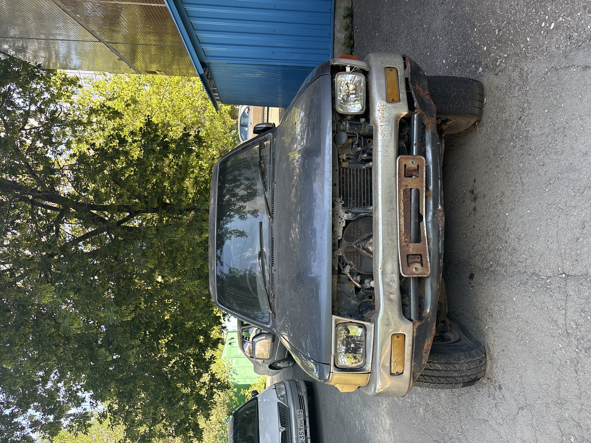 A weathered, dark gray SUV is parked on an asphalt surface. The front of the SUV is visible, showing a missing grille and radiator, and a rusted bumper with a license plate mount. The headlights are intact, but the passenger-side front tire appears to be flat. To the left of the SUV, a white sedan is parked, and behind it, a light-colored sedan is visible. A large, leafy tree with green foliage shades the front of the SUV. To the right of the SUV, a bright blue corrugated metal structure stands adjacent to a tan building with reflective windows. The scene is an outdoor urban environment, likely a parking lot or side street in Novosibirsk, Russia, given the license plate on the white sedan which reads "E351XH 154". The lighting suggests it is daytime with dappled sunlight filtering through the tree.