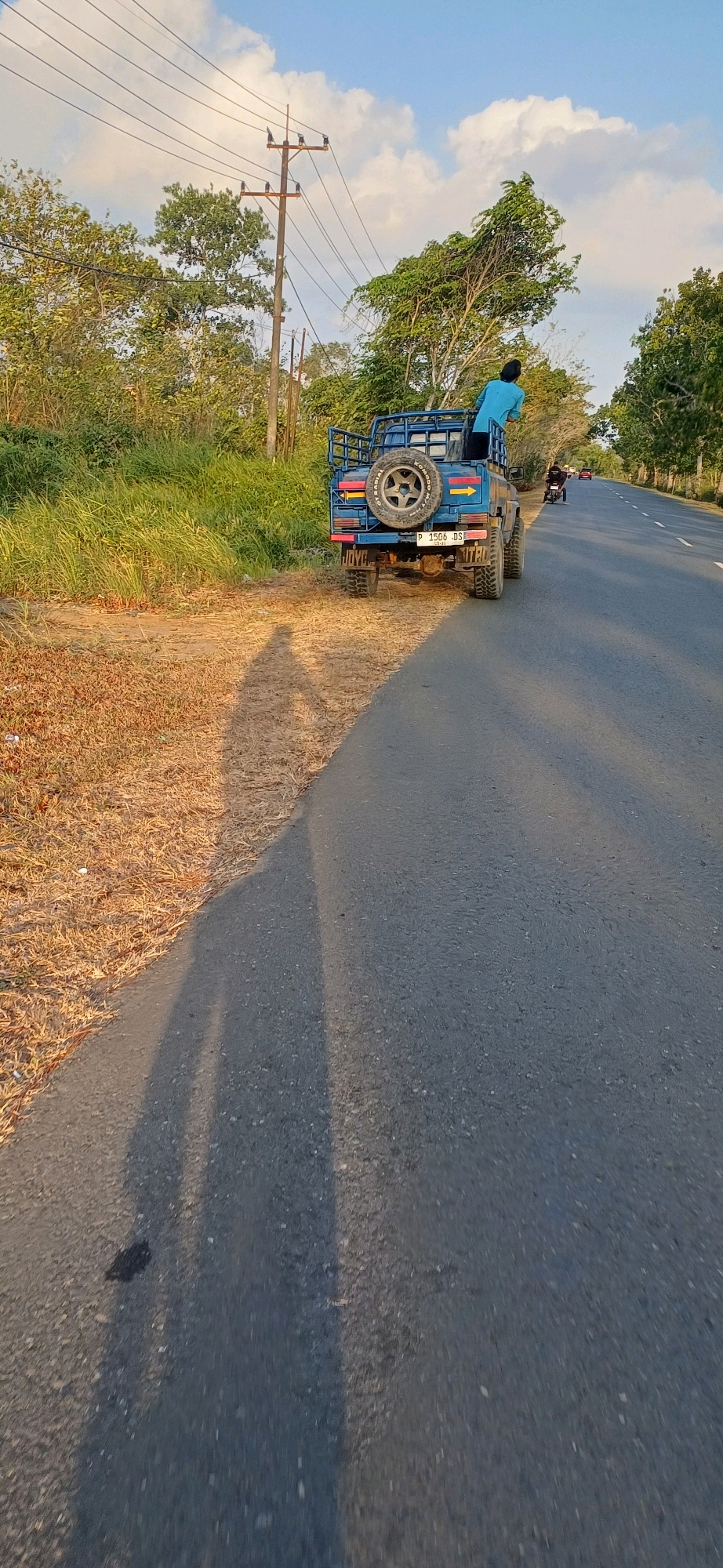 A blue Suzuki Jimny is parked on the side of a road in Kersik Putih, Indonesia. A person in a light blue shirt is standing in the back of the vehicle, looking towards the right. The vehicle has a spare tire mounted on the back, and a license plate that reads "P 1506 DS". The road is paved and has white lines indicating lanes. To the left of the road is dry grass and some trees. In the background, there are more trees and a power pole with wires. The sky is blue with some white clouds. The sun is setting, casting long shadows. The scene is peaceful and serene.