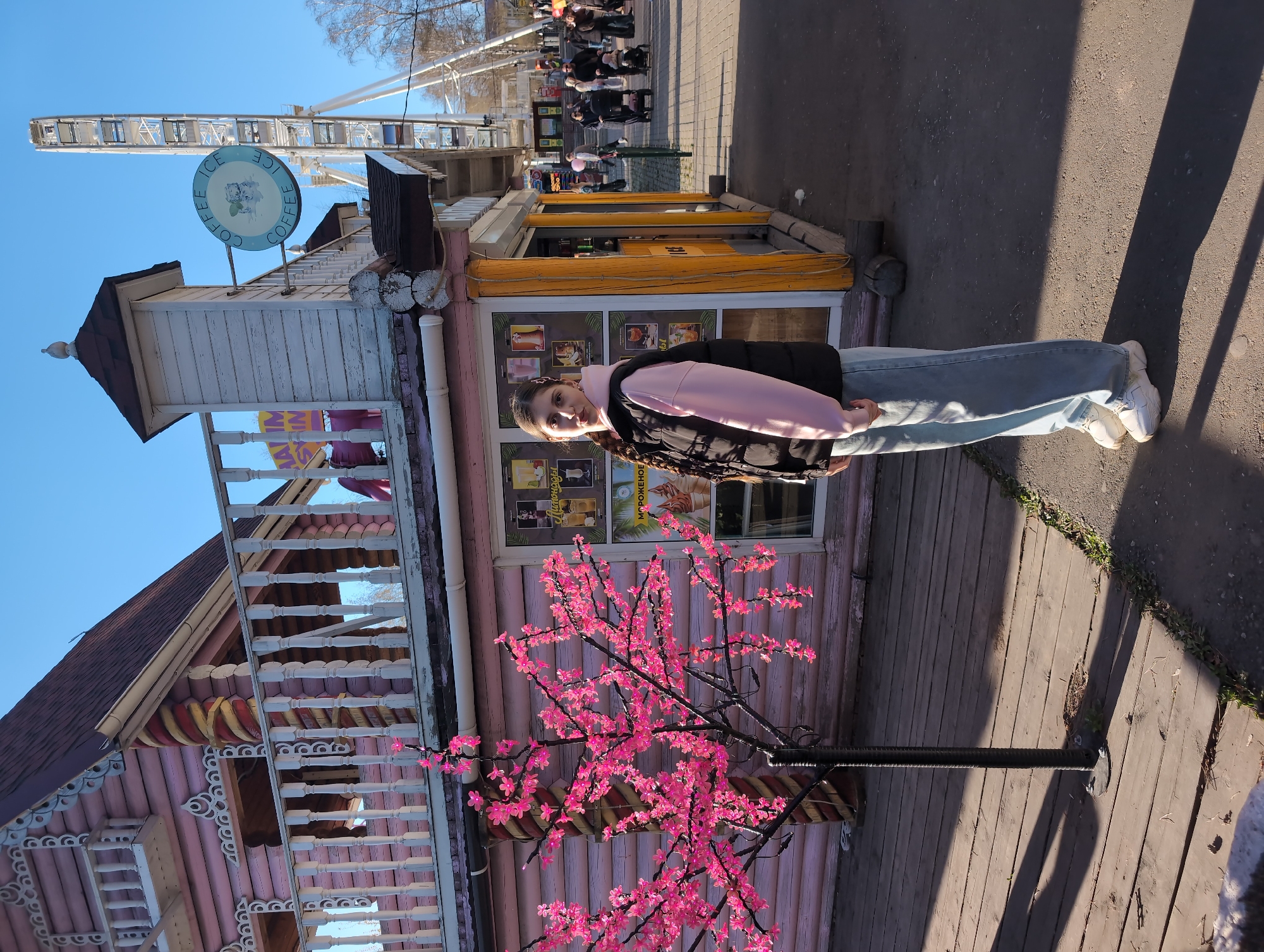A young woman stands next to a pink, ornate wooden building with a faux cherry blossom tree in Izhevsk, Russia. The building has a sign that reads "Ice Coffee Ice." In the background, a Ferris wheel is visible. A group of people walks along the sidewalk. The weather appears sunny.