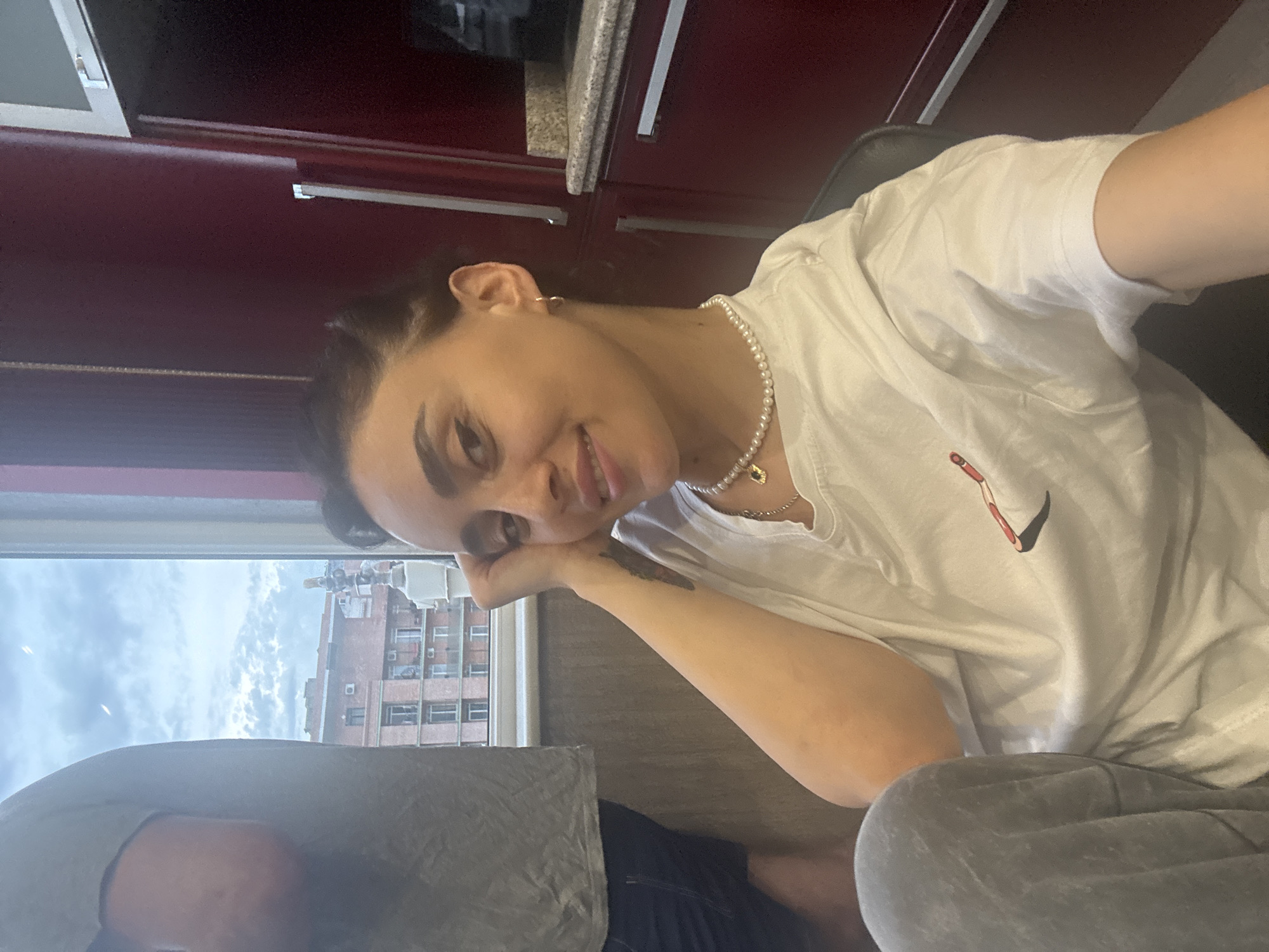 A young woman with dark hair pulled back is sitting and looking at the camera with a slight smile. She is wearing a white t-shirt with a small logo, a pearl necklace, and a gold hoop earring. Her left arm is resting on her face. In the background, a person in a grey t-shirt and dark shorts is visible, partially out of frame. The setting appears to be indoors, with a window showing buildings and a cloudy sky, consistent with Tbilisi, Georgia. Red cabinetry is visible to the right of the woman.