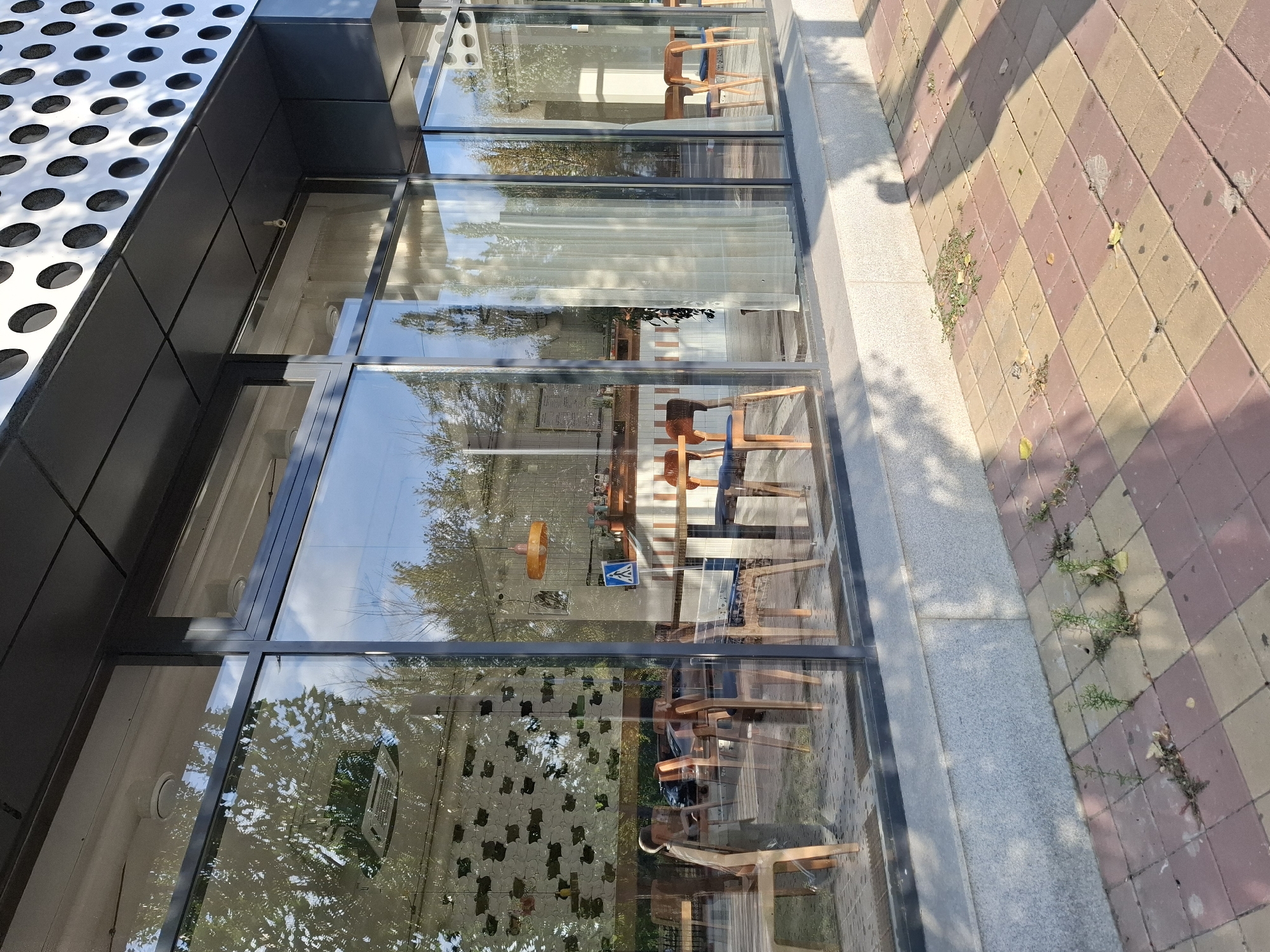 The image depicts the exterior of a modern building with large glass windows, located in the city of Lisakovsk, Kazakhstan. The building has a dark facade and a perforated metal accent on the upper section. Through the windows, the interior of what appears to be a cafe or restaurant is visible, furnished with wooden chairs and tables. Reflecting in the glass are trees and the sky. Outside, the ground is paved with a checkered pattern of light and dark pink square tiles, with some weeds growing between them. The lighting suggests it is daytime, with shadows cast across the pavement.