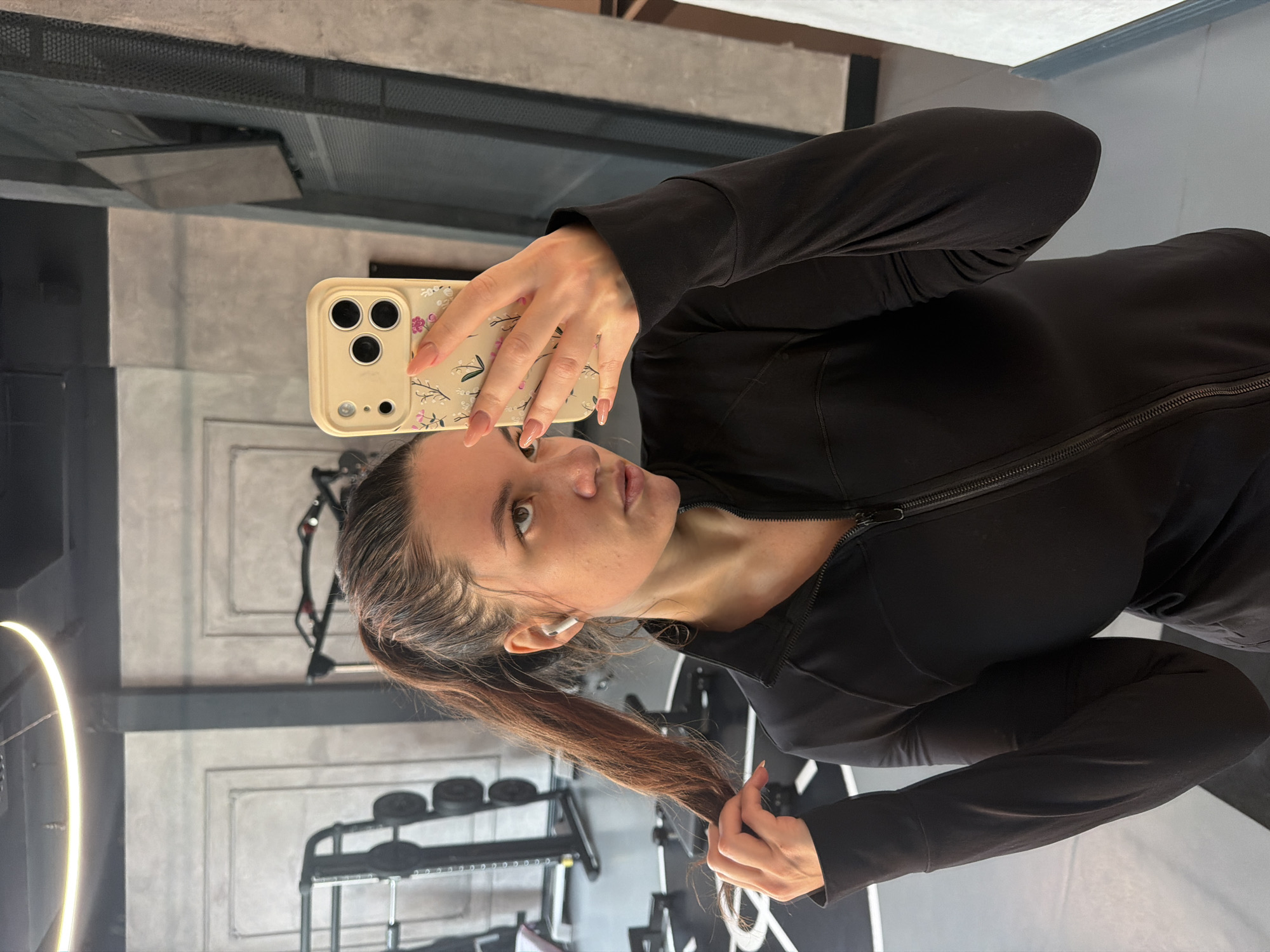 A woman with long brown hair, pulled back in a ponytail, is taking a selfie in what appears to be a gym. She is wearing a black long-sleeved jacket. In her left ear is a white wireless earbud. She is holding a cell phone with a light-colored floral case in her right hand. Behind her is gym equipment, including weight racks and exercise machines. The room has gray and white walls with black accents. The image orientation is rotated counter-clockwise. The city of Belgrade, Serbia is not discernable from this image.