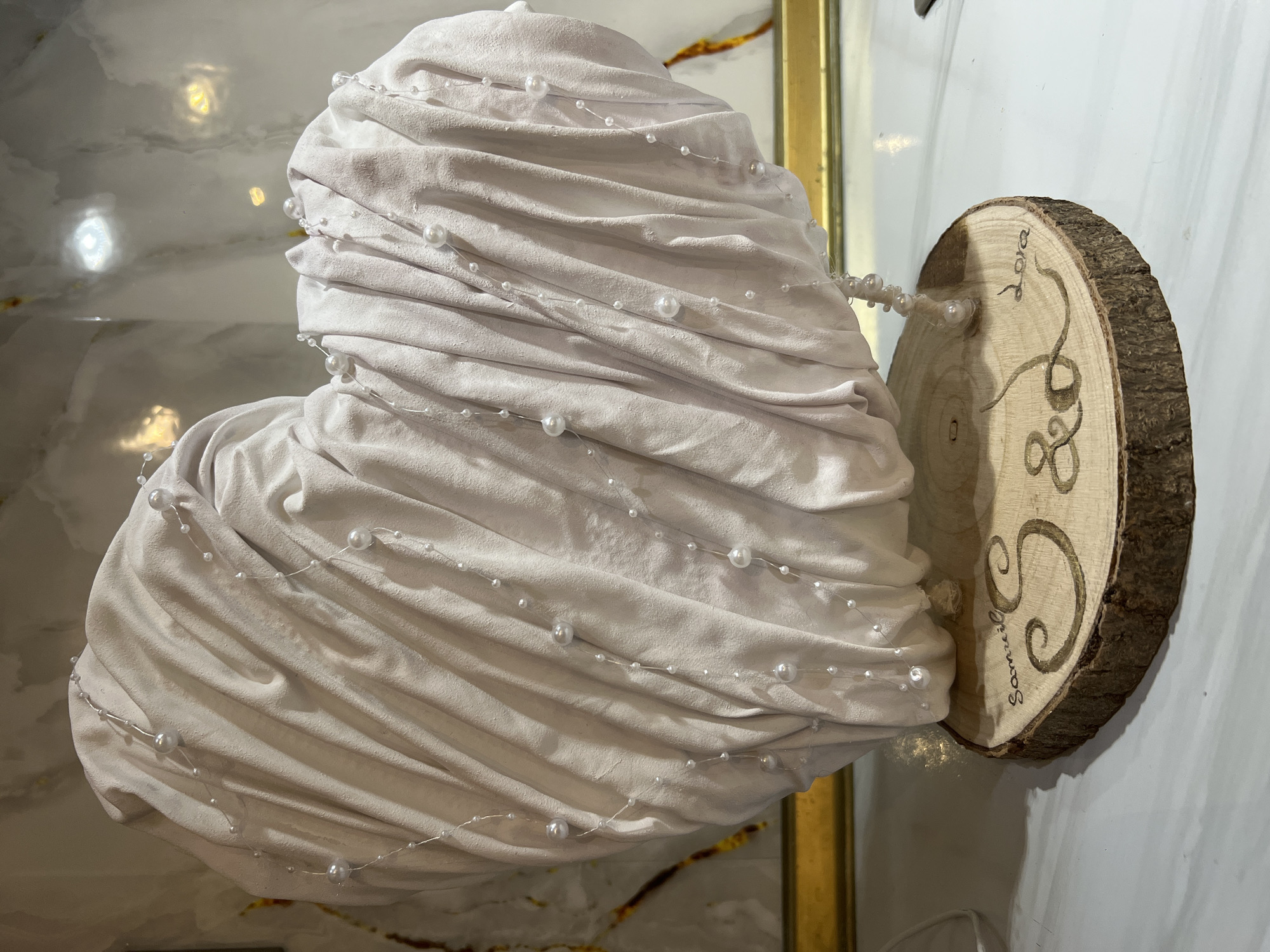 The image shows a decorative heart-shaped object made of draped white fabric adorned with strings of small white pearls. The heart is attached to a circular wooden slice with bark edges, on which the names "Samuel" and "2016" are inscribed. The background consists of what appears to be a marble or tiled surface and a section of a gold-colored frame or decorative trim. The picture provides no indication that it's in the city of Burgas, Bulgaria.