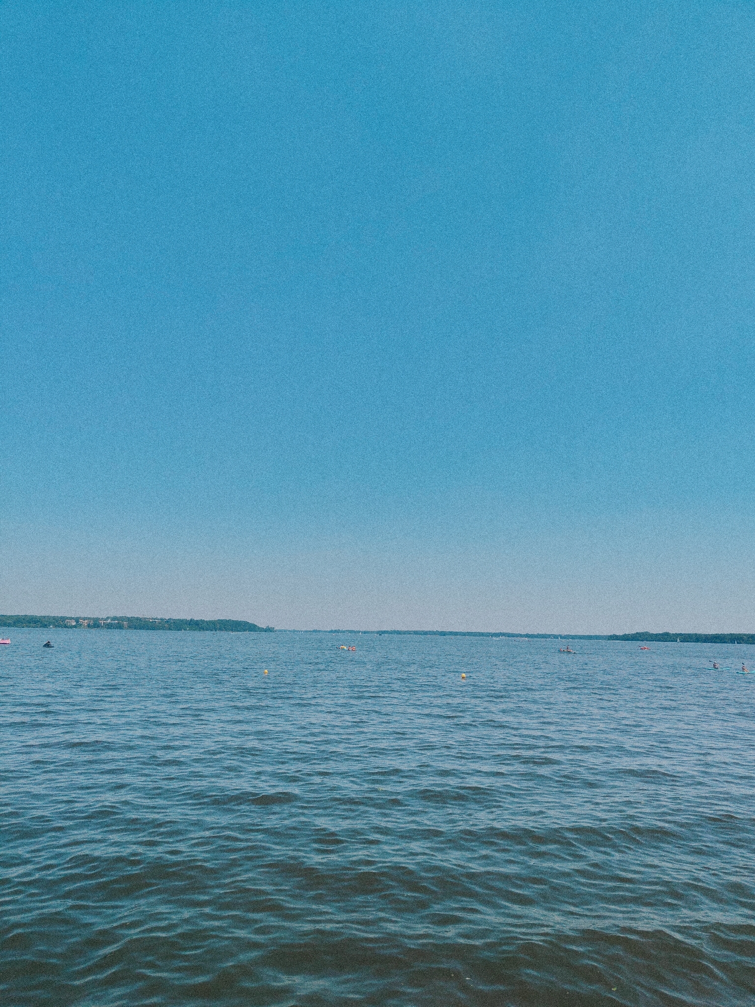 The image depicts a wide expanse of calm blue water under a clear, bright blue sky. In the distance, a tree-lined shore is visible, with some buildings nestled amongst the trees, suggesting an urban or suburban presence along the waterfront. Several small yellow buoys are scattered across the water, possibly marking a swimming or boating area. A few individuals are engaged in water activities: one person is on a stand-up paddleboard, another is in a kayak, and there are other distant figures in boats or on personal watercraft. The overall scene conveys a peaceful, recreational atmosphere on a body of water, likely a lake or a wide river. Given the description of Nieporęt, Poland, this location is likely the Zalew Zegrzynski (Zegrze Reservoir), a popular recreational area near Warsaw.