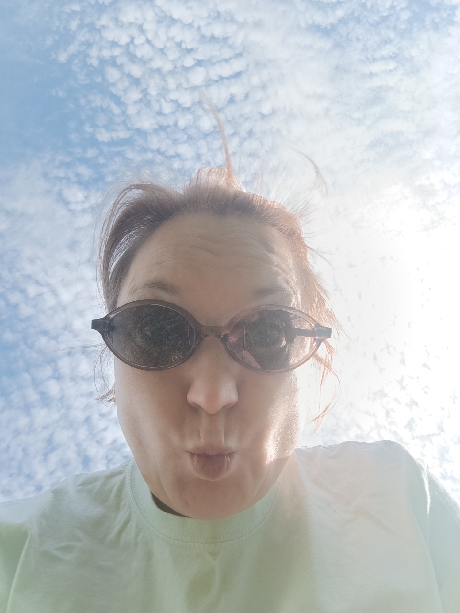 A woman with reddish-brown hair, styled with a few strands sticking upwards, is pictured from a low angle, looking down at the camera with pursed lips. She is wearing sunglasses that reflect a landscape of grass and trees. The background is a light blue sky with small, white clouds. She is wearing a light green t-shirt. The bright sunlight is partially obscuring details on the right side of the frame. This image does not contain information that would indicate that it was taken in the city of Dubovaya, Russia.