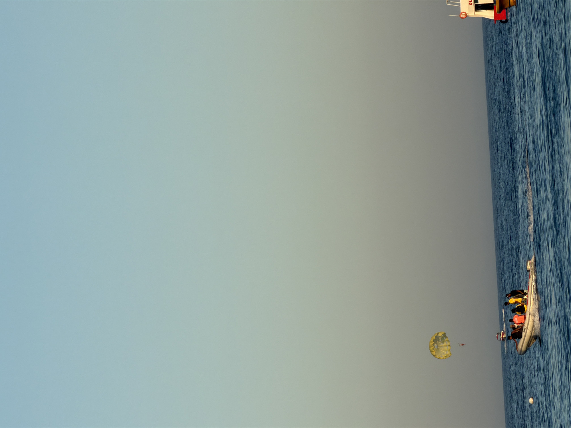 An image captures a wide expanse of the sea under a clear sky, characteristic of Sharm El Sheikh, Egypt. In the foreground, a motorboat navigates the blue waters, leaving a white wake. A group of individuals, some wearing bright orange and yellow life vests, are seated in the boat. Further in the distance, a yellow parasail is visible, with a person suspended beneath it, being towed by an unseen vehicle. To the right, a larger boat, possibly a ferry or tour boat, with white and red accents and the partial inscription "GL..." is seen moving away from the viewer. A white buoy floats on the water to the left of the motorboat. The overall scene depicts a typical day of water sports and marine activity in a popular tourist destination.