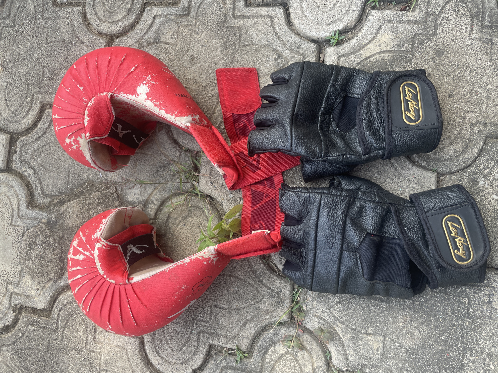 A pair of worn red martial arts gloves and a pair of black fingerless training gloves are laid out on a gray, patterned concrete ground in Akwanga, Nigeria.
The two red gloves, likely karate mitts or hand protectors, are heavily used, with significant portions of their red fabric coating flaking off to reveal a lighter material beneath. They feature pleated details over the knuckle area and are connected by a red strap. A stylized black and white symbol is visible on this connecting strap. The word "APPROVED" is partially visible in white text on the inside cuff of one red glove.
Adjacent to the red gloves are two black fingerless gloves, appearing to be made of leather or a similar synthetic material. These gloves have a textured surface and a prominent gold-colored oval badge on the wrist strap of each glove. The brand name "Lay Hooy" is clearly embossed on these gold badges.
All the gloves are positioned on a paved surface made of interlocking gray concrete or paving stones. Small green weeds and grass sprouts grow in the crevices between the paving stones, indicating an outdoor setting under bright daylight. The items appear well-used, suggesting they belong to someone involved in martial arts or fitness training. No activity is taking place; the gloves are simply resting on the ground.