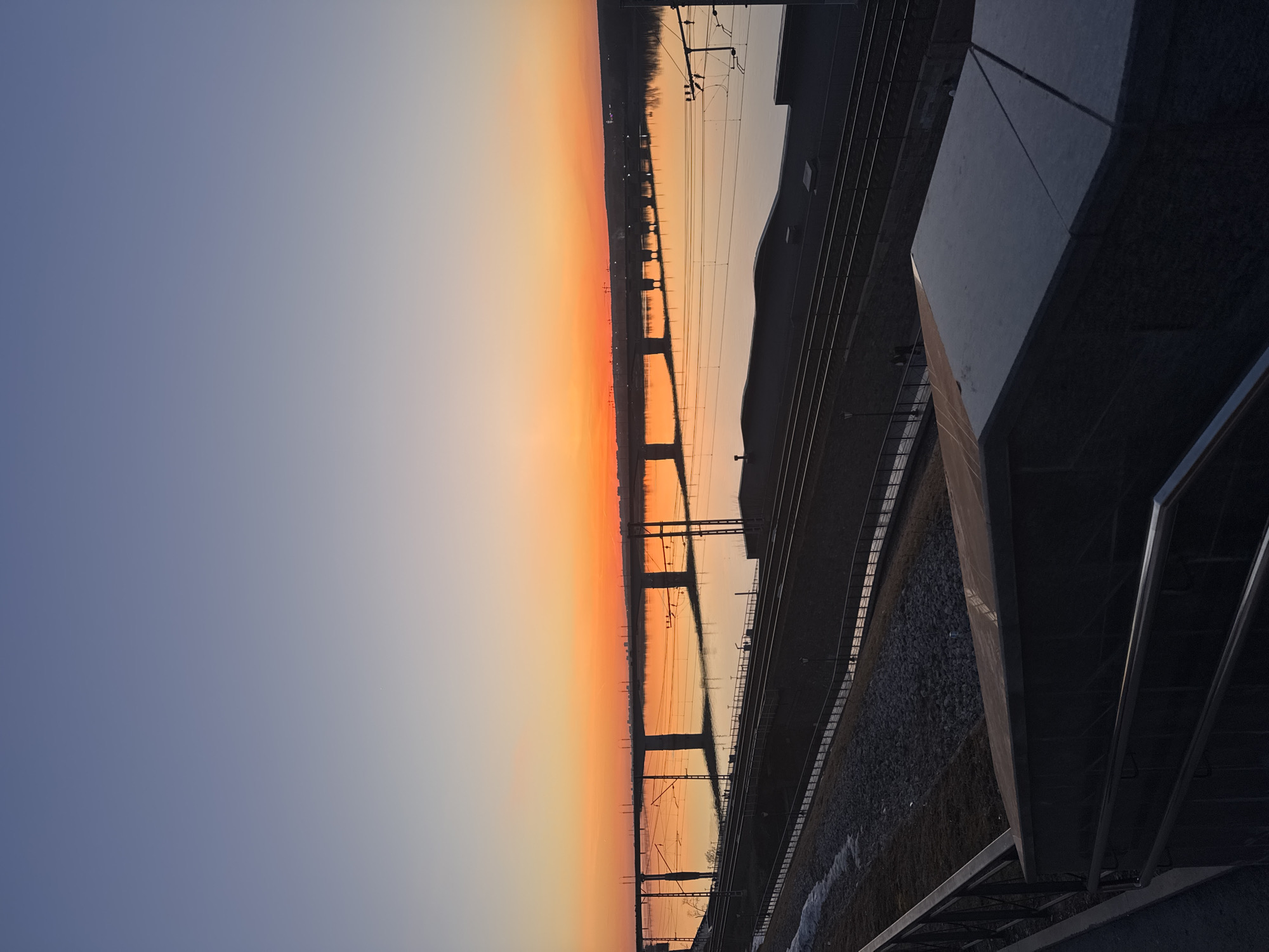 An eye-level, outdoor shot captures a railway bridge over the Kama River in Perm, Russia, at sunset. The sky is a gradient of pale blue to orange. The bridge's supports reflect in the water below. A railway track runs parallel to the riverbank, adjacent to a concrete structure and metal railings.