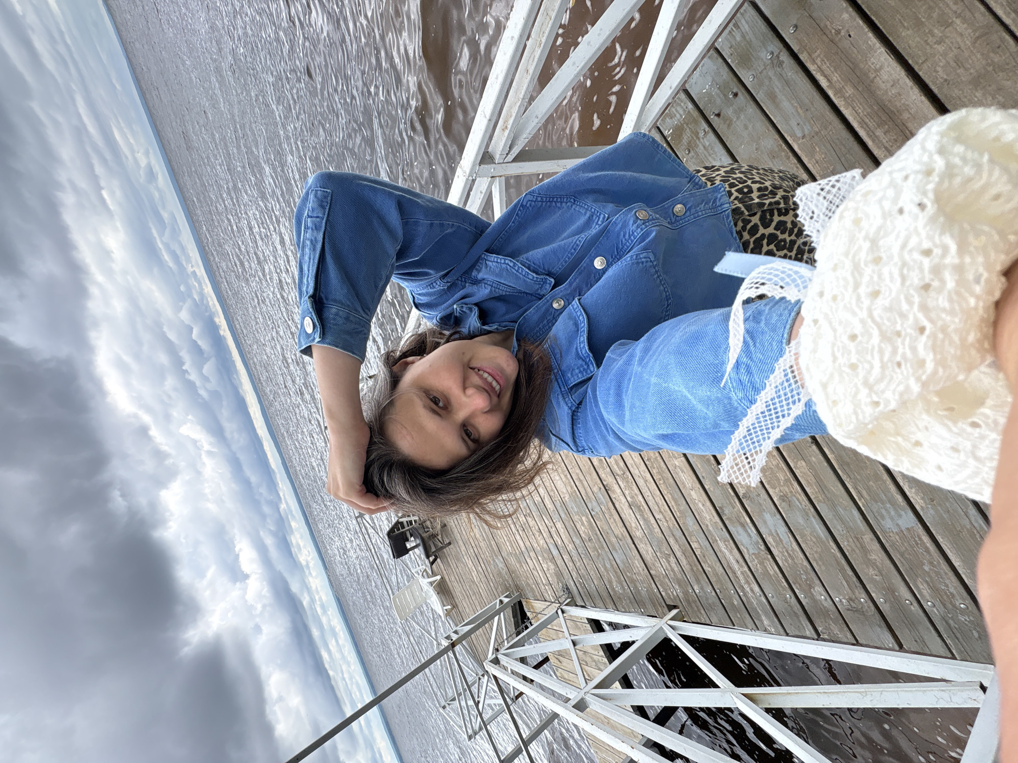 A young woman with dark hair is lying on a wooden dock, smiling at the camera. She is wearing a denim jacket and leopard print pants, and is holding a white, crocheted hat in her hand. The dock is on the water, and the water appears choppy. The sky is cloudy. The overall mood of the image is relaxed and happy. The location is Kostovaty, Russia.