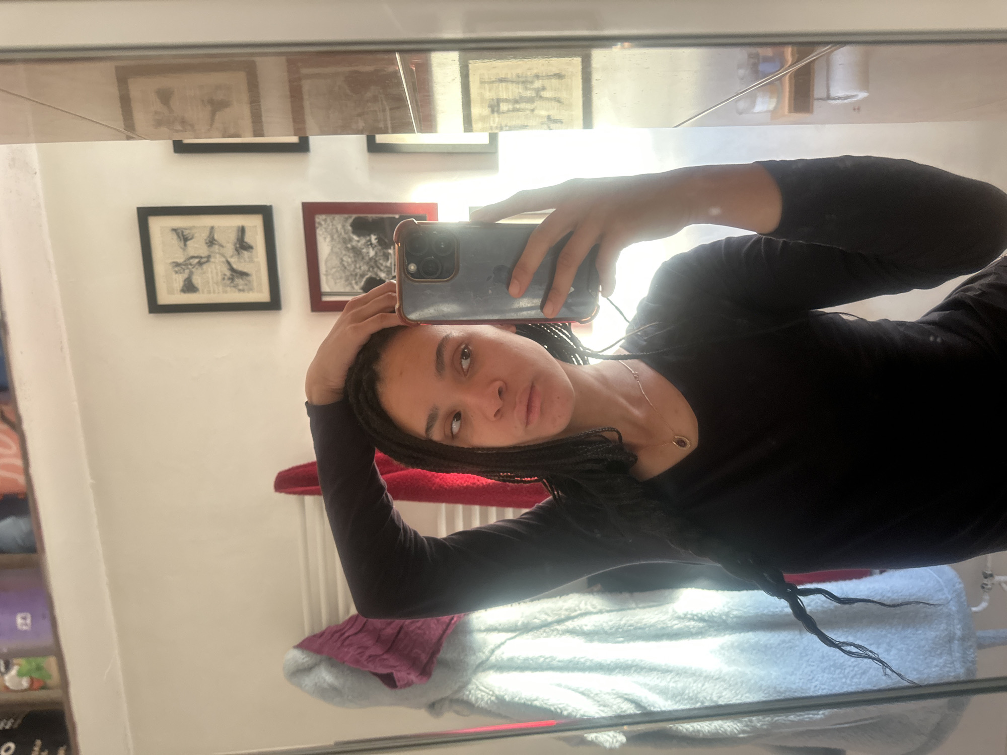 A young woman is depicted in a mirror selfie, holding a dark-colored smartphone with a clear case in her left hand, which is aimed at her reflection. Her right hand rests on her head. She has dark skin and long, dark braided hair. She is wearing a plain black long-sleeved top and a thin gold necklace with a small pendant. Her gaze is directed towards the camera with a neutral expression, showing a slight pout.
The setting is an indoor room, likely a personal space such as a bedroom or living area in the London Borough of Ealing, United Kingdom. The background walls are white, displaying several framed pictures. One prominent black-framed artwork features detailed black-and-white botanical or abstract illustrations on what appears to be aged paper. Another red-framed picture, partially visible, contains a black and white image. A strong, bright light source, likely a window, casts significant illumination from the right side of the frame, creating a glare on the mirror's surface. Below the woman's right arm, a red towel or fabric is draped, and a light blue fluffy blanket or towel is visible further down, alongside a darker purple fabric. A partially visible shelving unit with various items is reflected on the far left. The bright lighting suggests it is daytime. No discernible text is visible within the image.