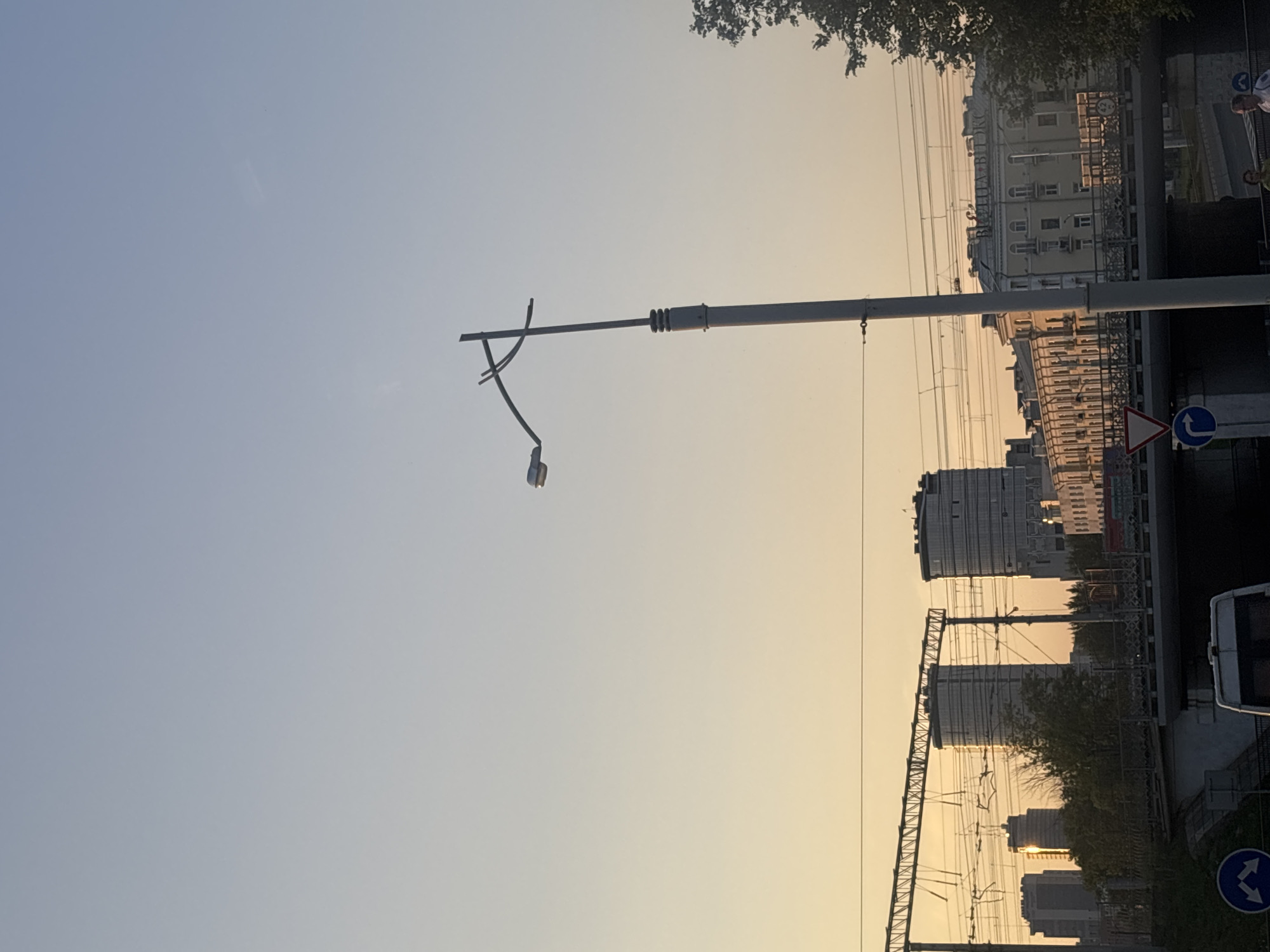 The image shows a city skyline at sunset, likely in Minsk, Belarus, given the presence of a building with Cyrillic lettering. In the foreground, a lamppost stands tall, its light fixture angled towards the left. Behind it, a railway bridge spans across the frame, with train tracks and overhead power lines visible. On the other side of the bridge, several buildings are scattered across the cityscape. Two prominent cylindrical buildings with glass facades dominate the mid-ground, while a collection of other structures, including a building with ornate detailing and arched windows, are visible further in the distance. The sky transitions from a pale blue at the top to a warm, golden hue near the horizon, indicating the time of day is sunset. A tree with dark green foliage is partially visible on the right side of the image. Several cars and pedestrians are present near the bridge, suggesting a busy urban environment.