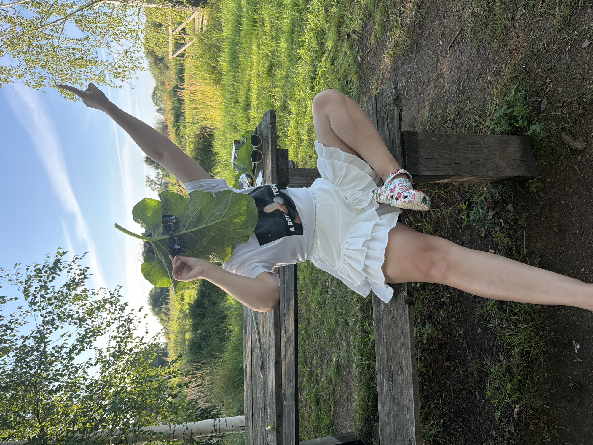 A woman sits on a weathered wooden picnic bench in a grassy field with trees in the background, holding a large green leaf in front of her face. She is wearing a white t-shirt with a graphic print and a white ruffled skirt. Her right arm is extended upwards, and she is wearing a floral-patterned shoe. Sunglasses rest on the picnic table next to her. The scene appears to be outdoors, possibly in a park or rural area. The location is identified as Dubrova, Russia.