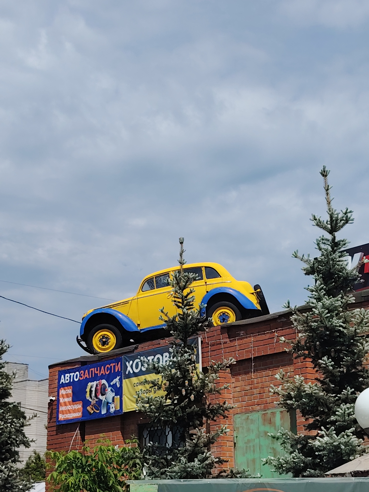 A yellow vintage car with blue accents is perched atop a brick building, its front tires visible and angled upwards. The building has a red brick facade, with what appears to be Christmas lights strung across the top section. Below the car, a large blue banner with text in Russian and images of car parts is displayed, with another yellow banner to its right. Several conifer trees, some of which appear to be dusted with snow or frost, are situated in front of the building. A large, slightly rusted green garage door is visible on the right side of the building. In the background, a multi-story white building is partially visible. The sky above is overcast with a pale blue hue. The scene is set against a backdrop of a cloudy sky.