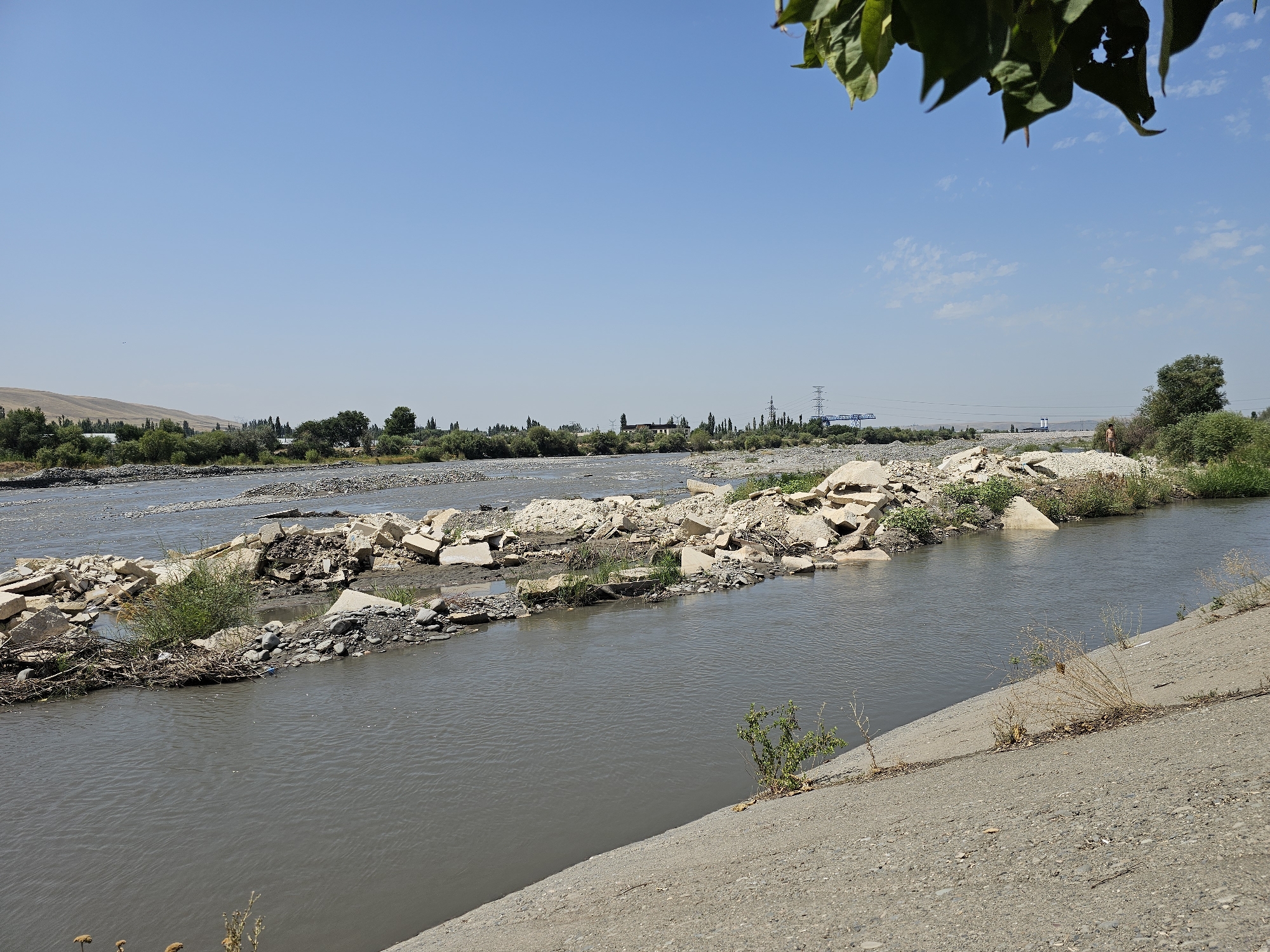 A wide, murky river flows through a landscape of arid hills and sparse vegetation. The river is partially obstructed by a debris dam composed of large, broken concrete slabs, rocks, and branches. On the right bank, a concrete embankment slopes down to the water, covered in gravel and dry brush. In the distance, a power line tower and some buildings are visible, hinting at human presence. The sky is a clear, pale blue with a few wispy clouds. The image appears to be taken on a sunny day. The scene depicts a river that has been impacted by debris and possibly floodwaters, creating an unnatural obstruction within the natural flow of the water.