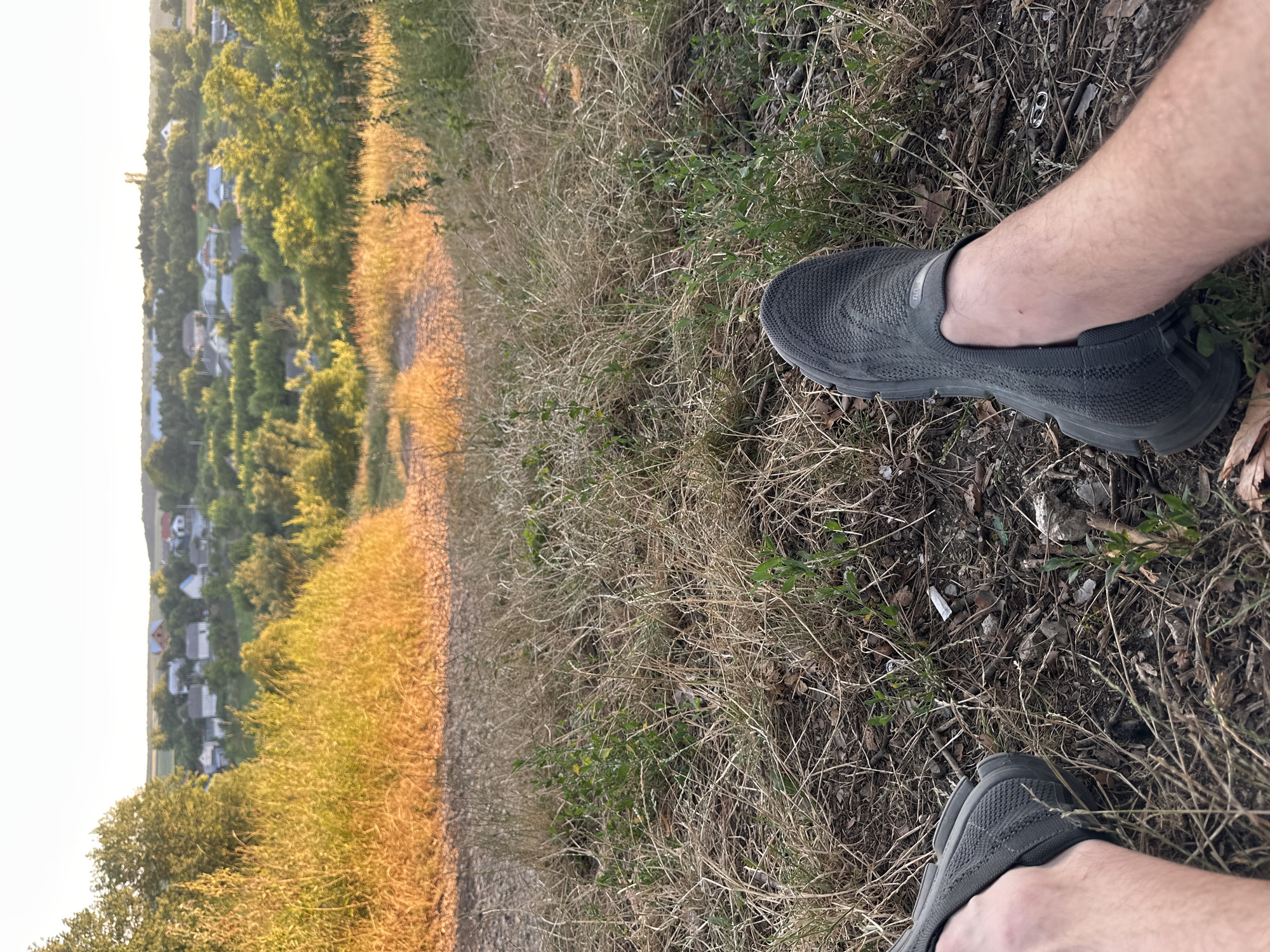 The image captures a point-of-view perspective of a person sitting on dry grass, facing a gentle slope that leads down to a village. In the foreground, the lower legs and feet of the person are visible, clad in dark gray slip-on athletic shoes. The ground is covered in a thick layer of dry, brown grass, interspersed with small green plants and scattered debris.
The slope is characterized by more dry grass and a dusty path winding down. Beyond the slope, a small village is spread out, with numerous houses featuring a variety of roof colors, including red, blue, and white. These houses are nestled amidst lush green trees and vegetation, suggesting a rural or semi-rural setting. The background consists of a hazy sky with the soft glow of what appears to be late afternoon or early evening sunlight. The overall scene suggests a peaceful moment of rest in the outskirts of Makiv, Ukraine, overlooking the local community.