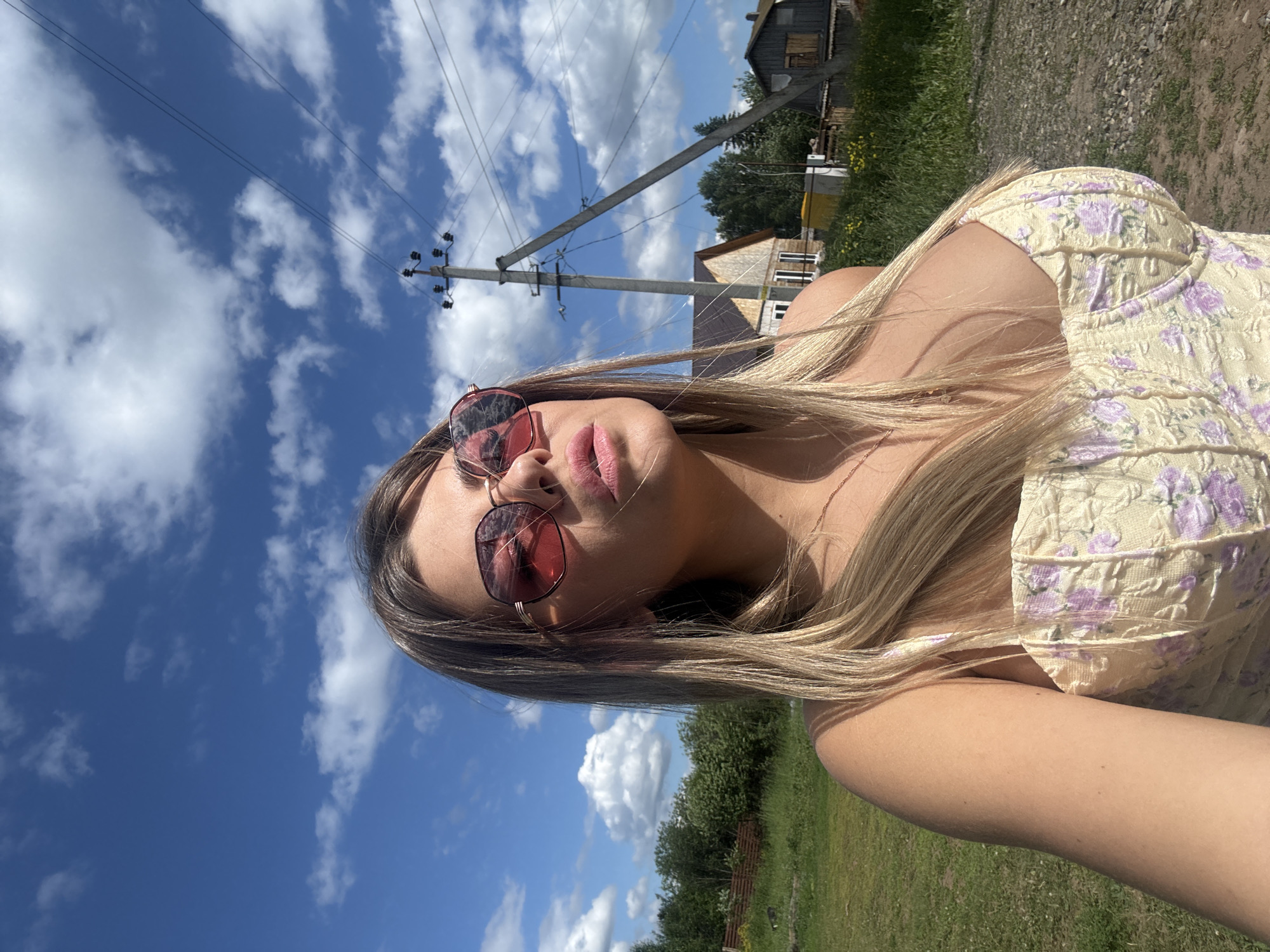 A fair-skinned woman with blonde hair and pink sunglasses is captured in a low-angle, outdoor selfie. She is wearing a light yellow corset-style top with a floral pattern in shades of purple and pink. The background features a bright blue sky with scattered white clouds, a utility pole with wires, some trees and foliage, and a few buildings, suggesting a rural or semi-rural setting. The overall impression is one of a sunny day in a village. Based on the provided information, this scene is located in the village of Pastukhovo, Russia.