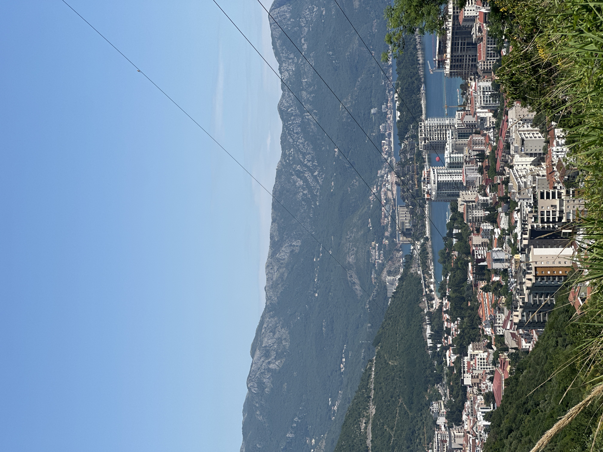 The image shows a view of the city of Podostrog, Montenegro, situated between a mountain and a body of water. The sky is clear and blue. Mountains are visible on the left side of the image, their slopes covered in greenery. Overhead, power lines stretch across the sky. The city itself consists of a mix of residential buildings, hotels, and other structures, densely packed along the waterfront and stretching up the slopes of the adjacent hills. The coastline is visible in the center of the frame, with water separating the urban area from the mountains. Green foliage is visible in the foreground, obscuring some of the view.