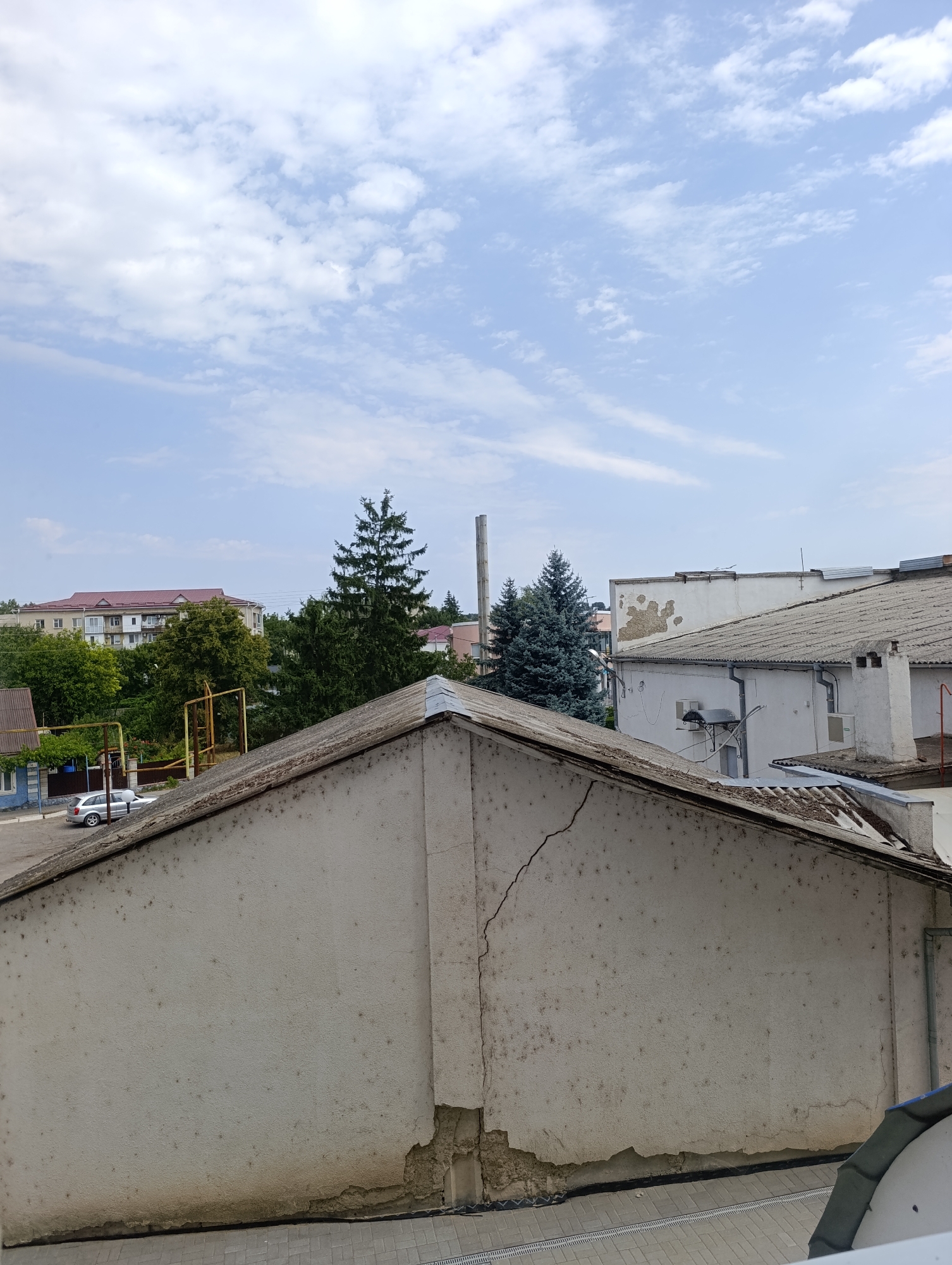 The image shows a view of buildings and trees under a cloudy sky in Dubăsari town council, Moldova. In the foreground, there is a white building with a damaged wall, revealing exposed bricks and a crack running across the surface. Beyond this building, a silver car is parked on a paved area next to a low blue building. Further back, several green trees are visible, including two tall, dark green conifer trees. Behind the trees, a multi-story building with a red roof stands, along with other buildings and a tall industrial chimney. The sky is a light blue with scattered white clouds.