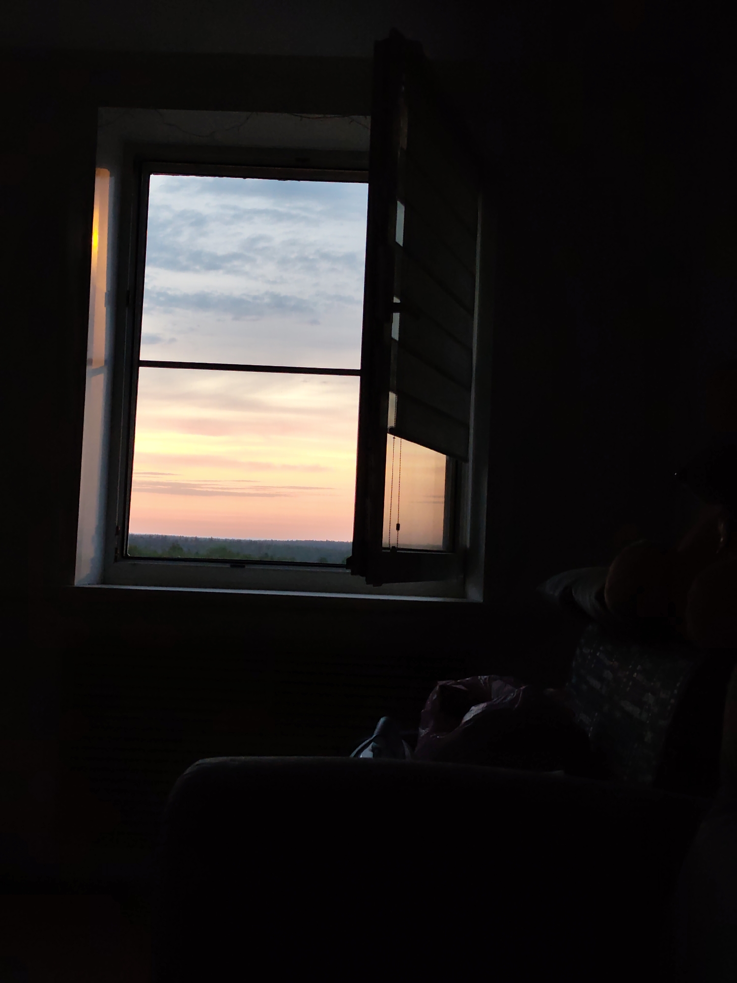 From a dimly lit room, a window reveals a view of the horizon. The sky is a blend of pale blues, pinks, and oranges, suggesting either sunrise or sunset. Below the sky, a dark strip of land, possibly trees or a distant landscape, is visible. The window itself is partially open, with one shutter extended outward, casting shadows within the room. The room is mostly dark, with only a suggestion of furniture and other objects discernible in the foreground. Given the location of Gremyachinsk, Russia, the landscape seen through the window is likely representative of that region.