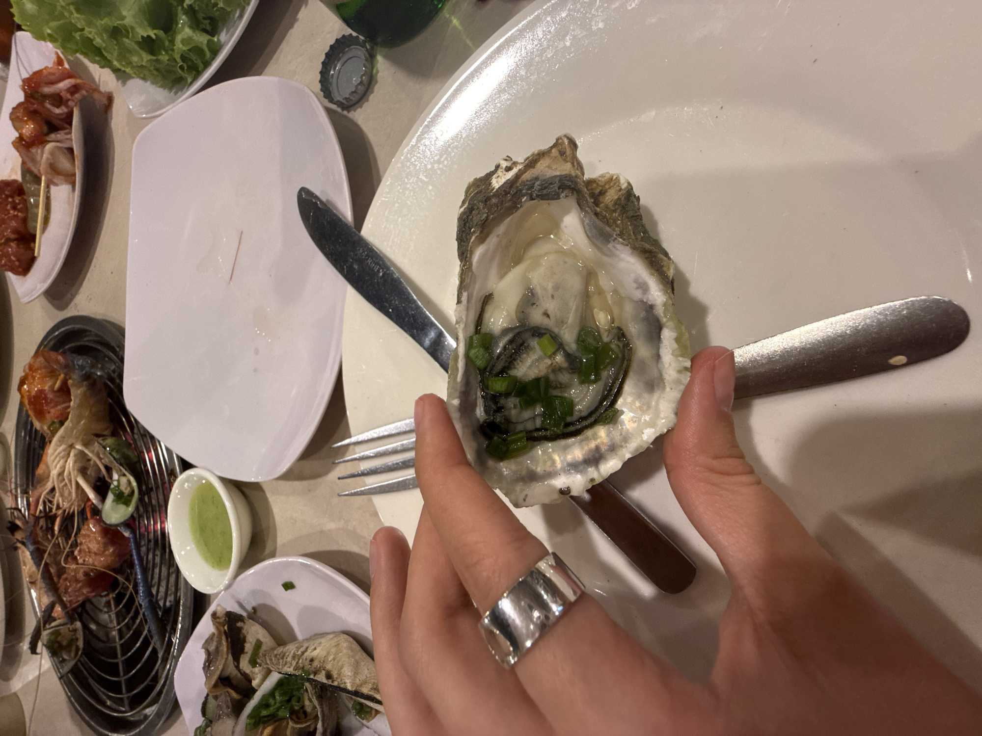 The image captures a close-up view of a person's hand holding a freshly shucked oyster, garnished with finely chopped green onions, against a backdrop of a dining table. The setting is likely an evening meal at a restaurant in Nha Trang, Vietnam, given the type of food and ambient lighting.
The person's hand, adorned with a wide, reflective silver-toned ring on the ring finger, securely grasps the oyster shell. The oyster inside is plump and glistening with its natural liquor. To the right, a metallic fork and knife are placed on a large, white rectangular plate. Another white plate holding fresh green lettuce leaves is visible in the upper-left corner, while a separate small white plate contains skewers of what appears to be kimchi or marinated meat.
To the left, a circular metal grill is laden with several large, cooked prawns or shrimp, along with other grilled seafood, suggesting a BBQ or shared cooking experience. A small white bowl filled with a vibrant green dipping sauce sits near the grill. In the lower-right, another plate with more oysters or similar shellfish can be seen. A metal bottle cap and a portion of a green bottle are visible in the background, implying beverages are part of the meal. The overall scene depicts a casual yet appealing seafood dining experience. No visible text is clearly discernible in the image.