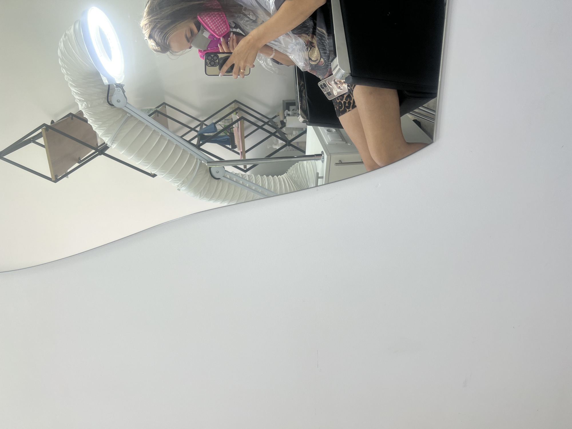 A woman is reflected in a mirror, seated in a chair, wearing a pink mask and holding a phone. She appears to be in a salon, with a large flexible lamp and ventilation system visible behind her. The environment is brightly lit and appears clean and modern.