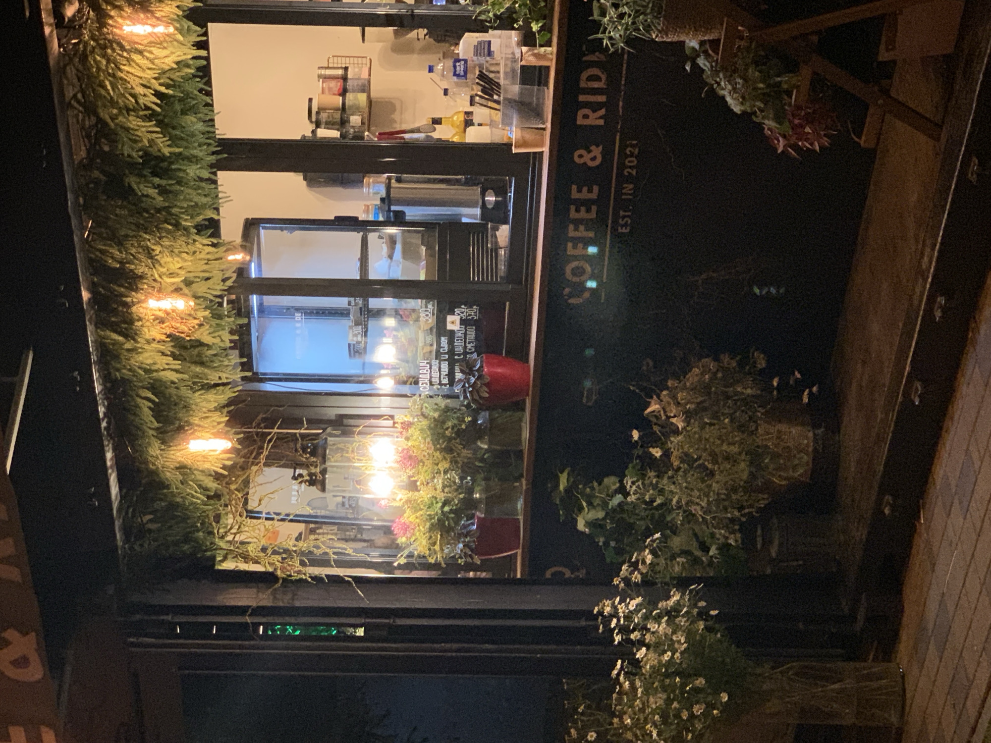 A coffee shop, identified by the signage "COFFEE & RIDE EST. IN 2021", is depicted at night. The shop has an open-air counter with a large window display showcasing various items. Inside the window, a refrigerated display case, coffee brewing equipment, and shelves stocked with jars and containers are visible. Outside, decorative plants and flowers are placed on the counter and in front of the shop. Incandescent lights and string lights illuminate the scene. The ground in front of the shop is paved with cobblestones. The overall atmosphere suggests a cozy and inviting establishment.